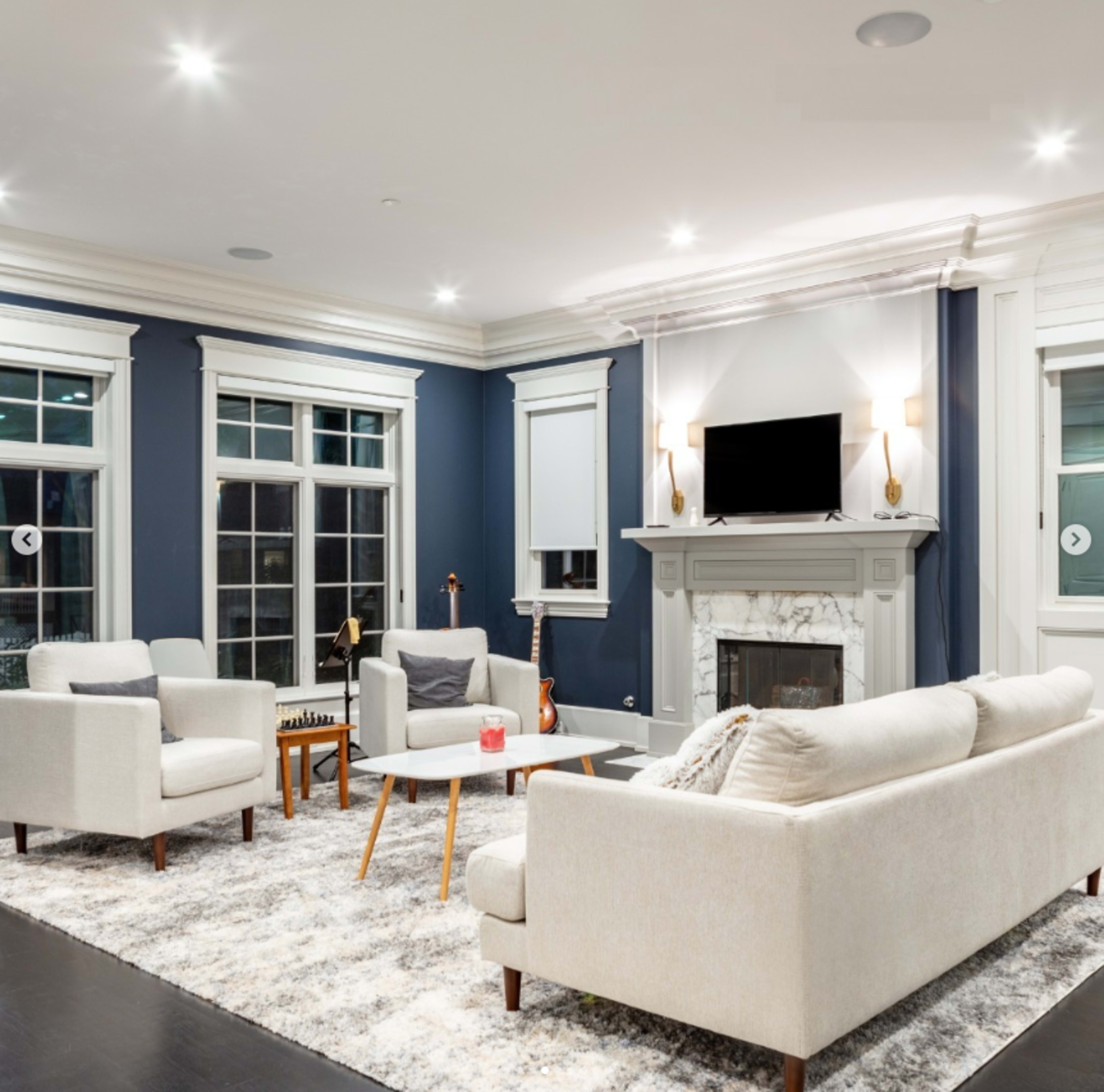 A modern living room features two light armchairs and a sofa arranged around a patterned rug, with a fireplace and a wall-mounted TV as focal points.