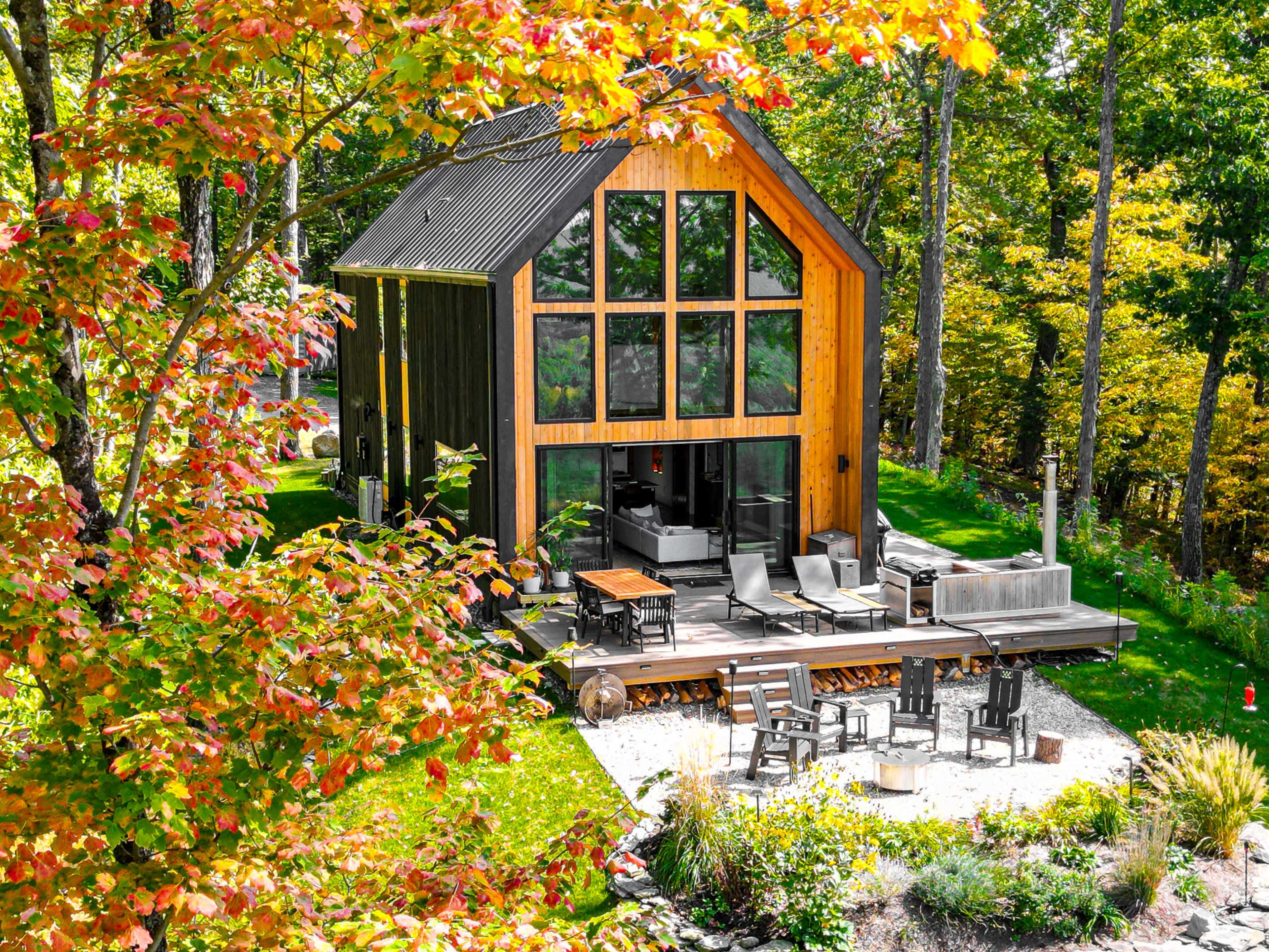 A modern cabin with large windows, surrounded by trees displaying fall foliage, features a patio area with outdoor seating and a fire pit.