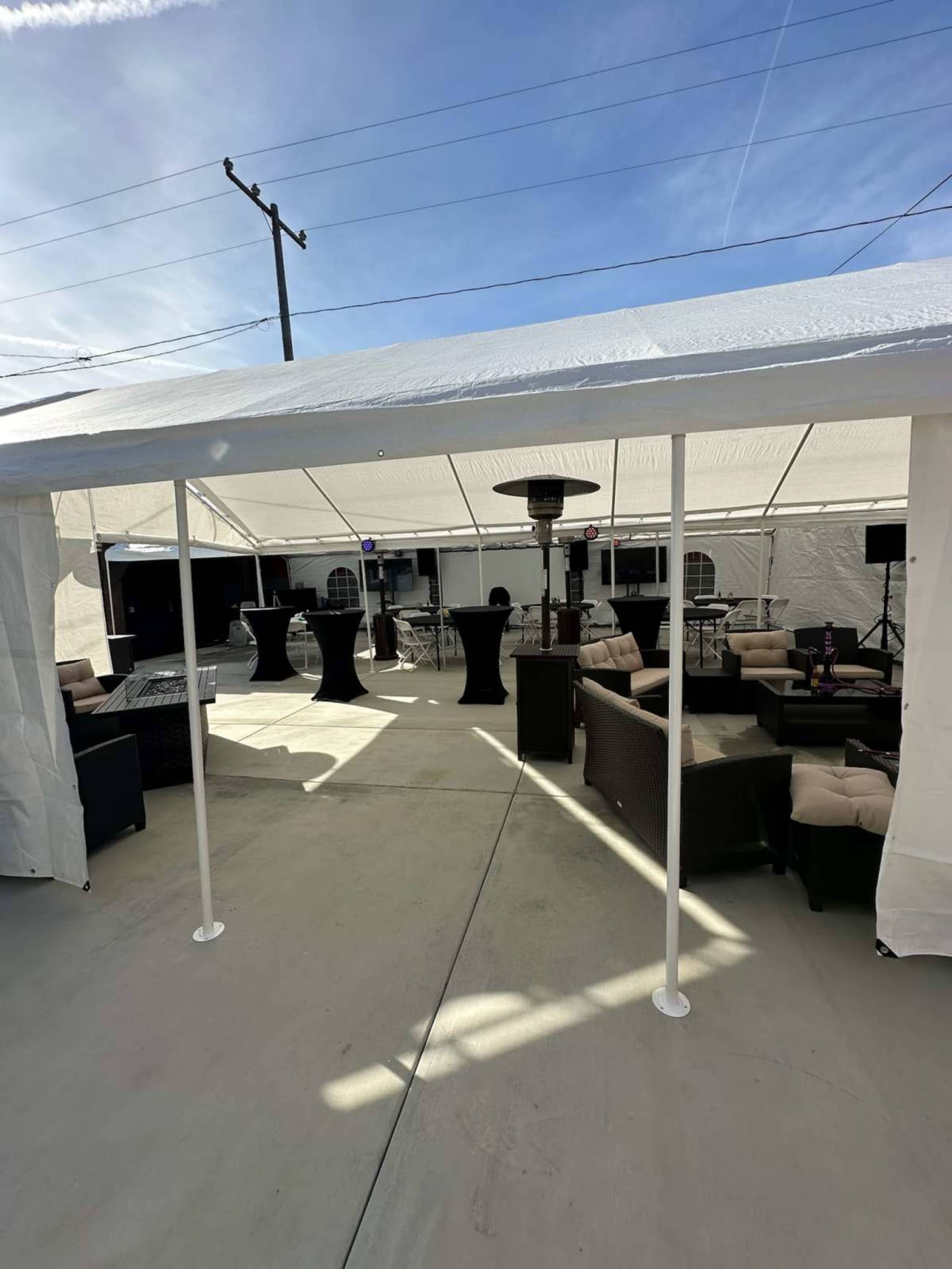 A white tent houses a seating area with wicker furniture and tall tables, situated on a concrete surface under a clear blue sky.