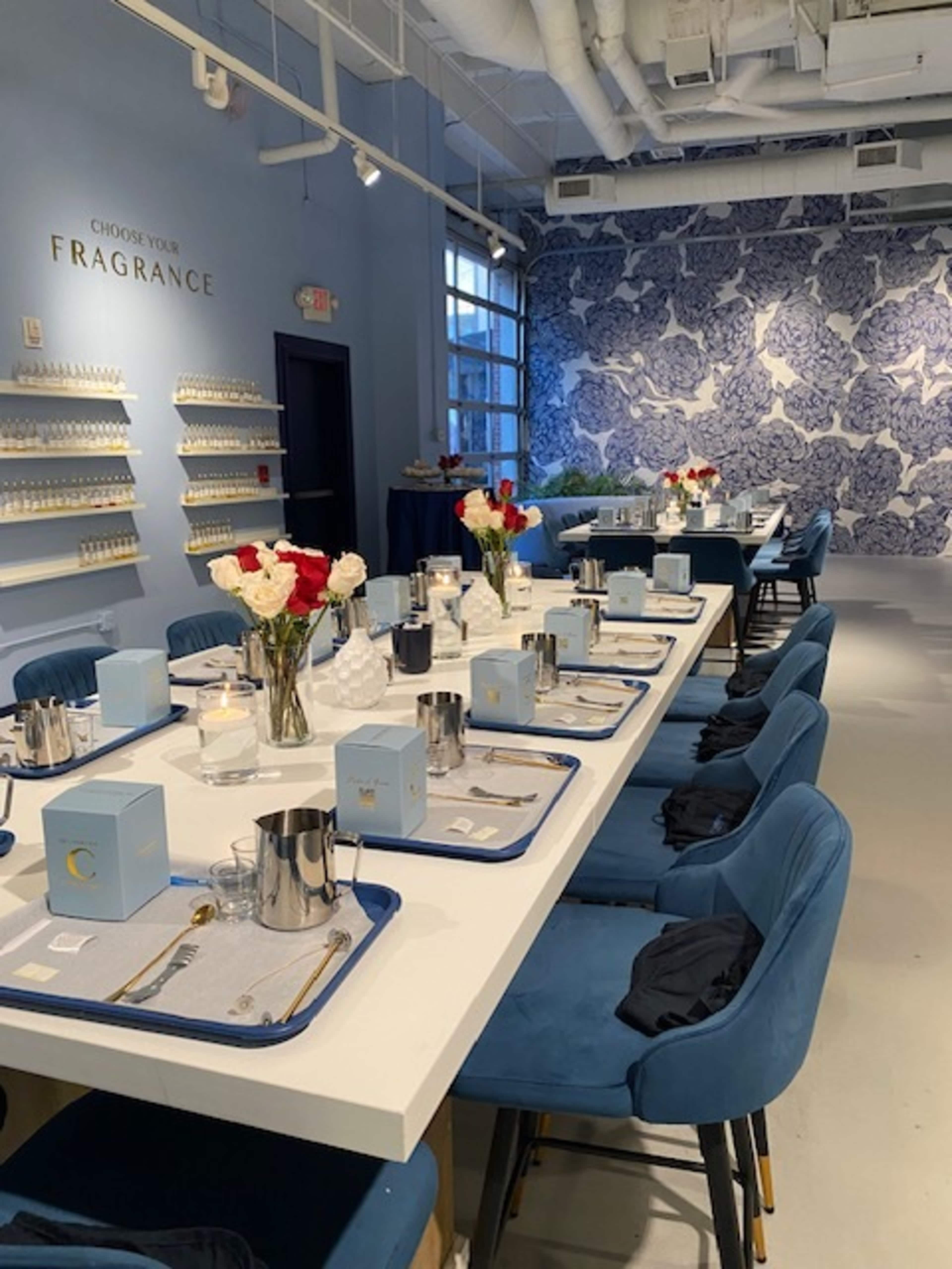 The image shows a modern, well-lit event space with a long dining table set with flower arrangements, trays, and boxes, against a decorative blue and white wall.