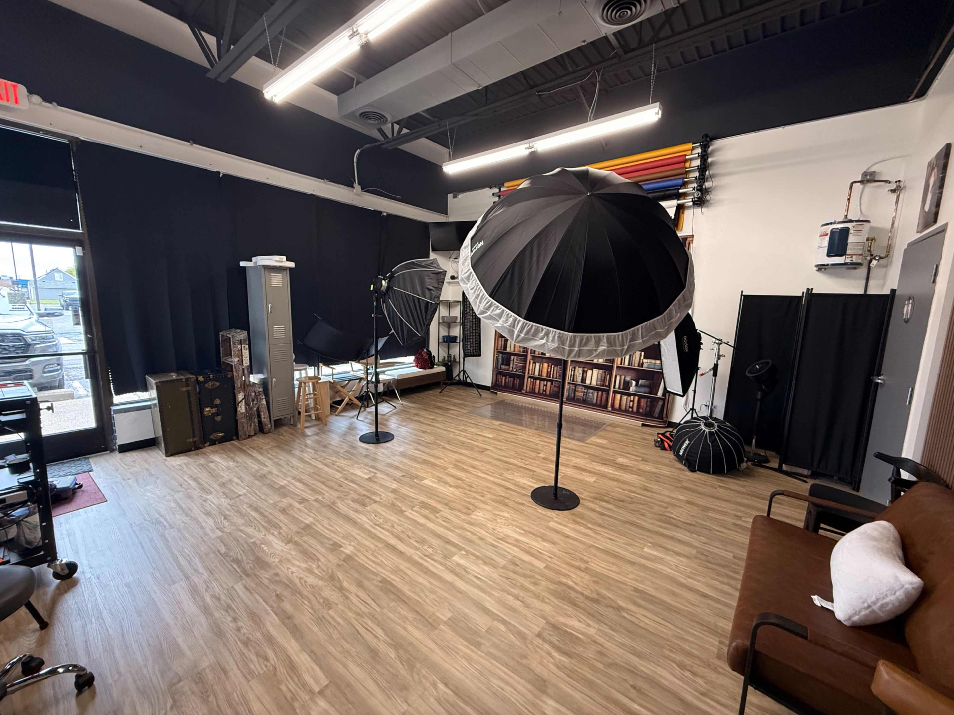 The image shows a photography studio featuring various lighting equipment, including large black umbrellas, a wooden chair, bookshelves, and a brown couch against the wall.
