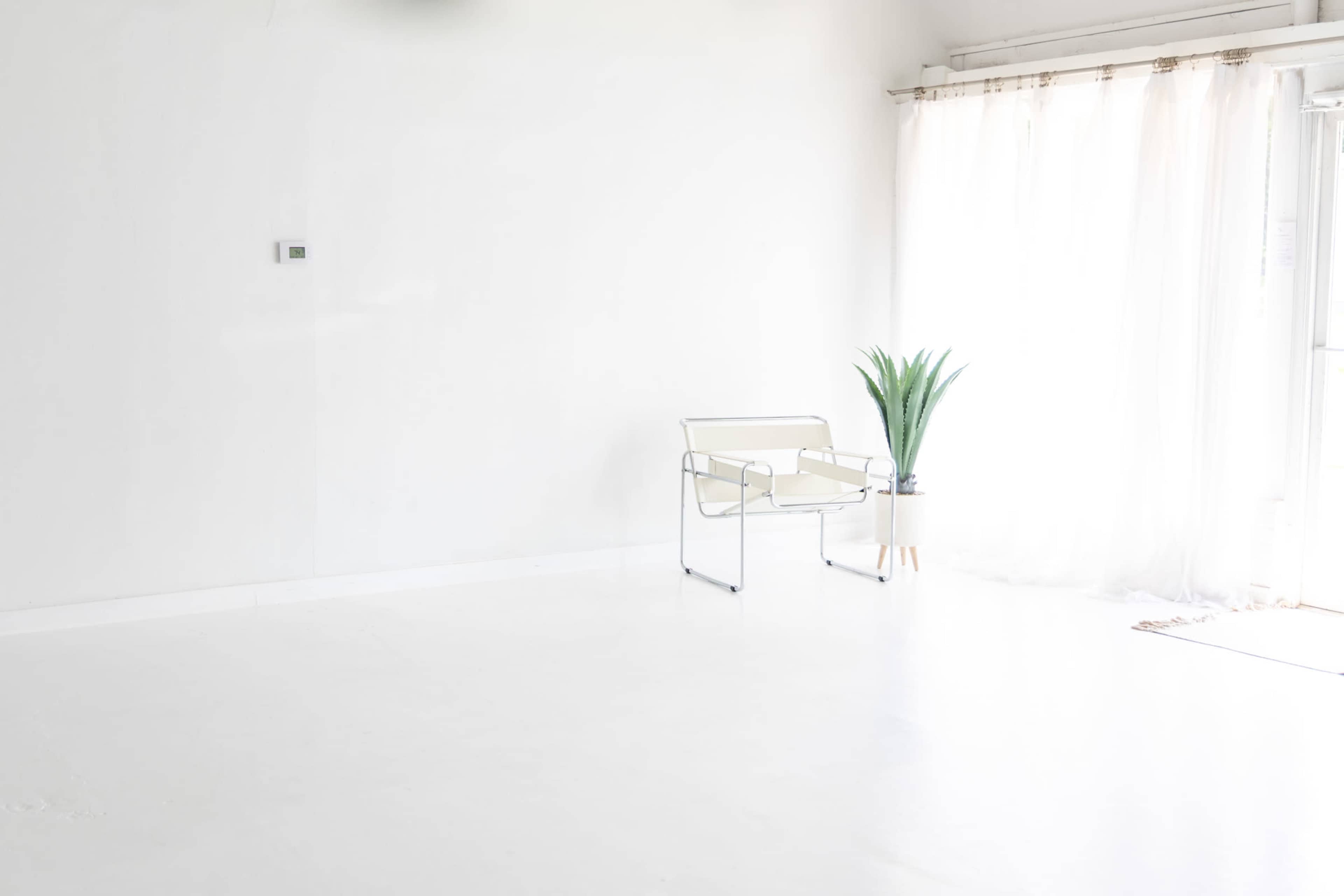 The image shows a minimalistic, empty room with a white floor, a single chair, a potted plant, and sheer curtains by a window.