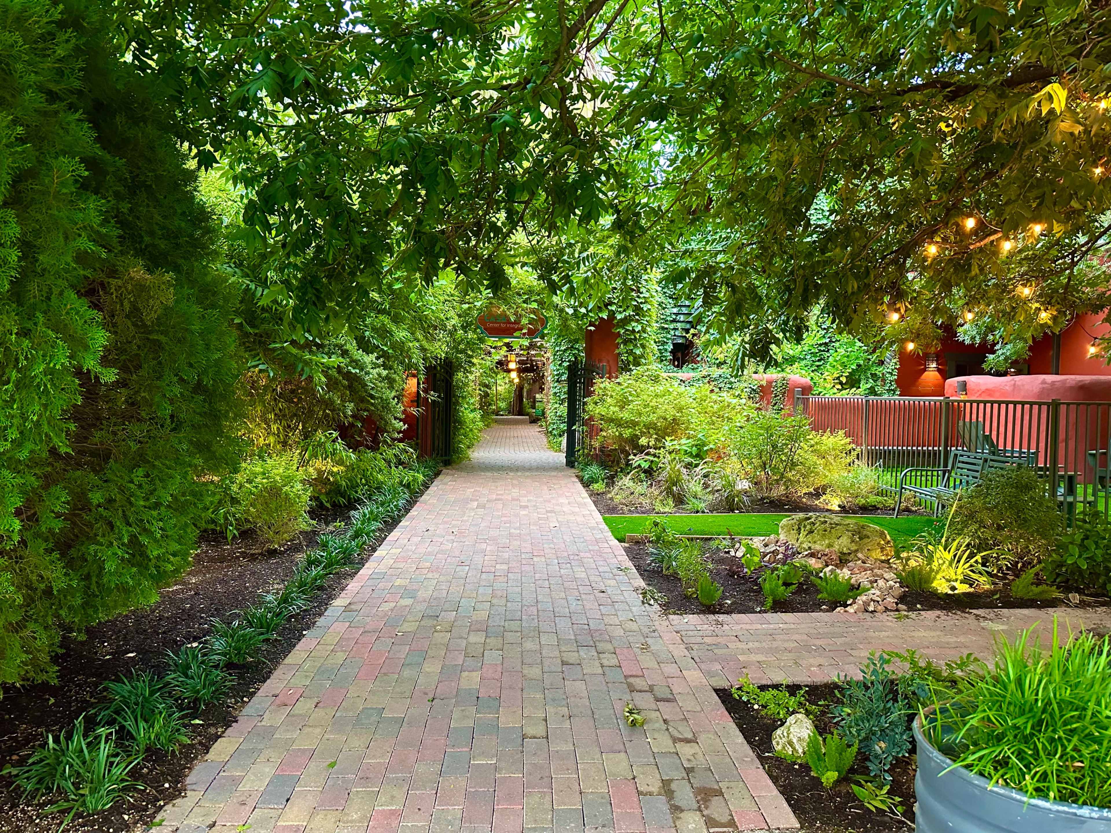 A brick pathway lined with greenery leads through a landscaped garden bordered by trees and decorative plants.