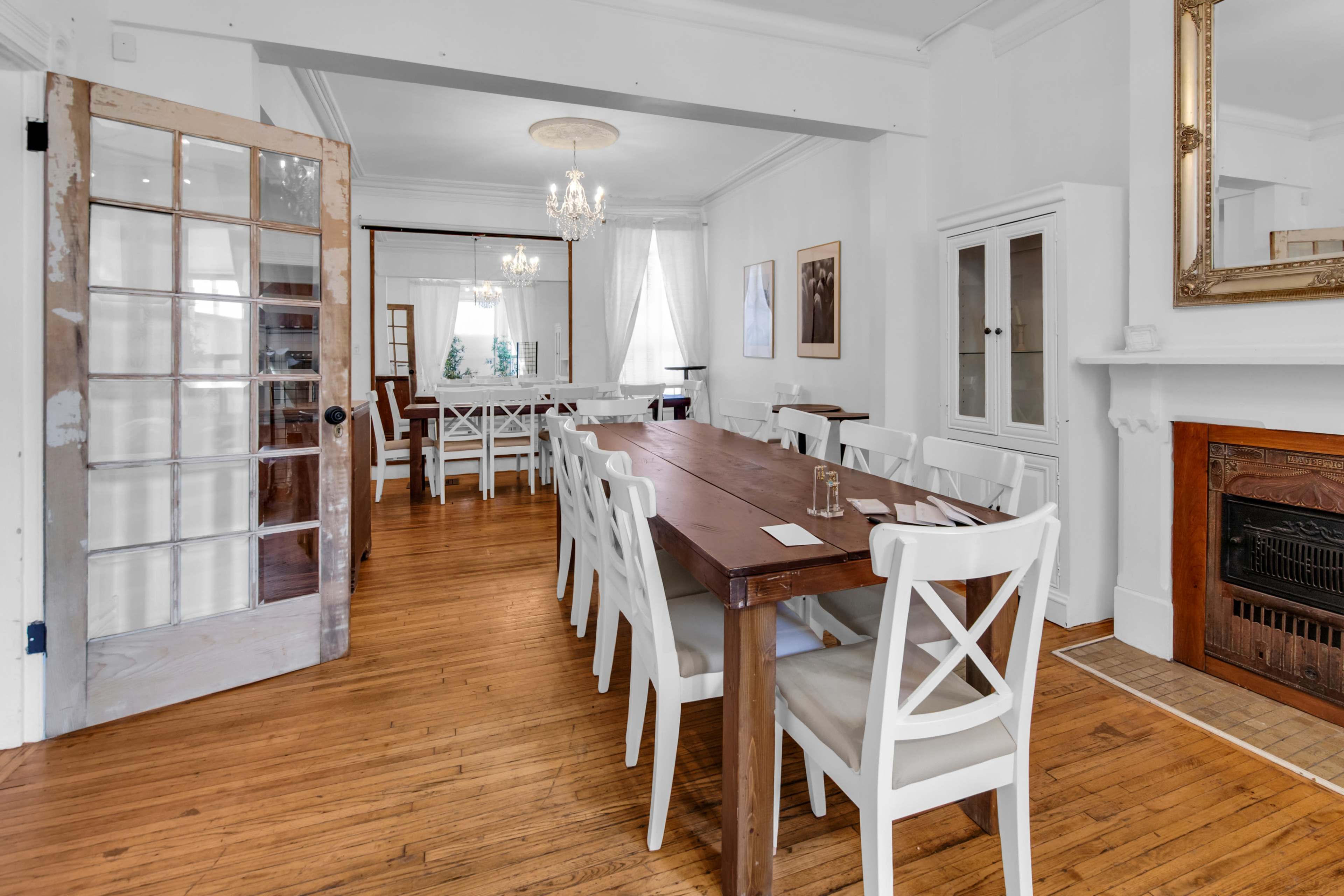A long wooden dining table with white chairs is positioned in a bright room that features large windows, a chandelier, and a decorative fireplace.