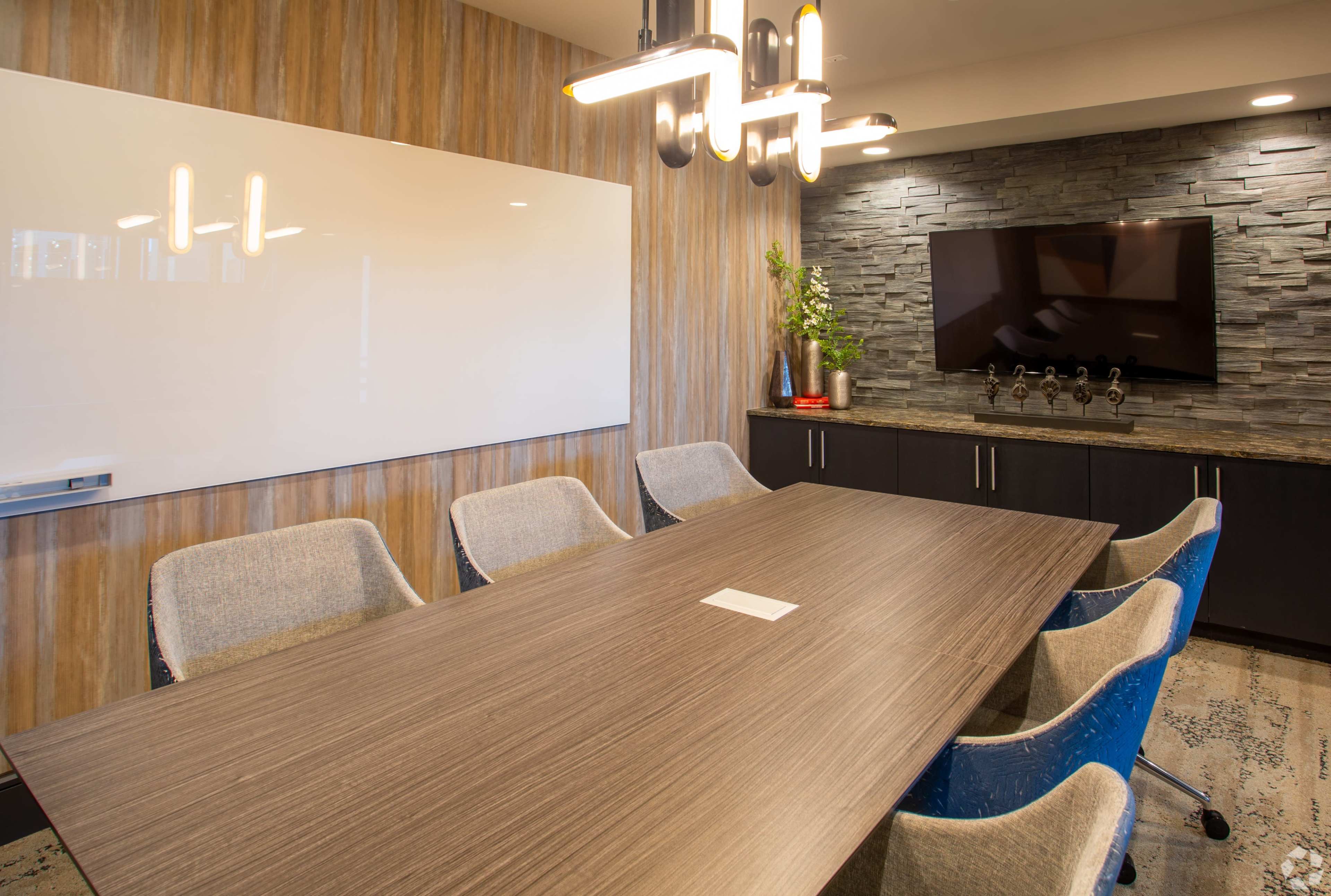 The image shows a modern conference room featuring a long wooden table surrounded by upholstered chairs, a whiteboard, a stone accent wall, and a television mounted on the wall.