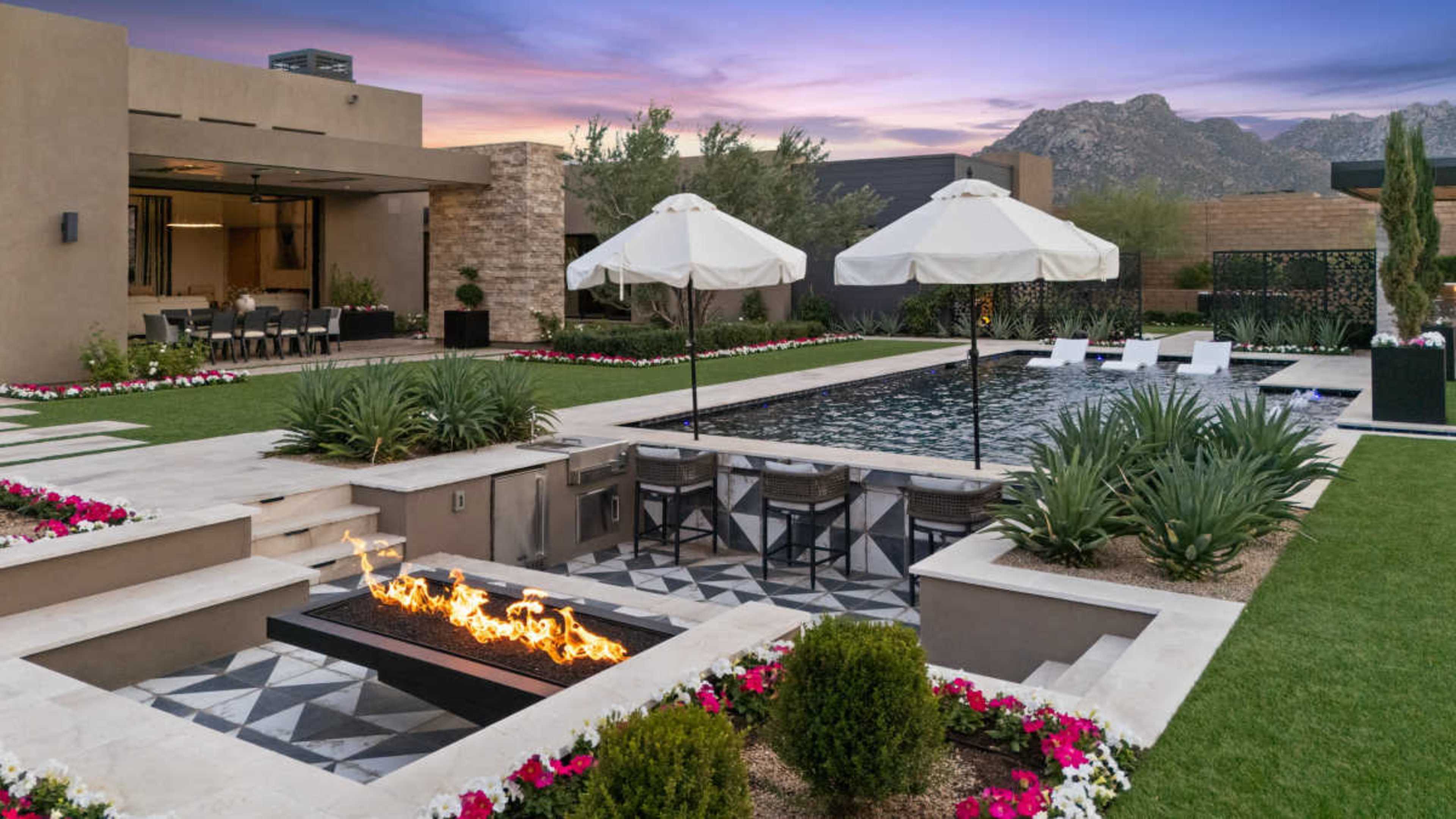 The image shows a modern backyard with a swimming pool, lounging areas under white umbrellas, and a fire feature, framed by landscaped gardens and mountains in the background.
