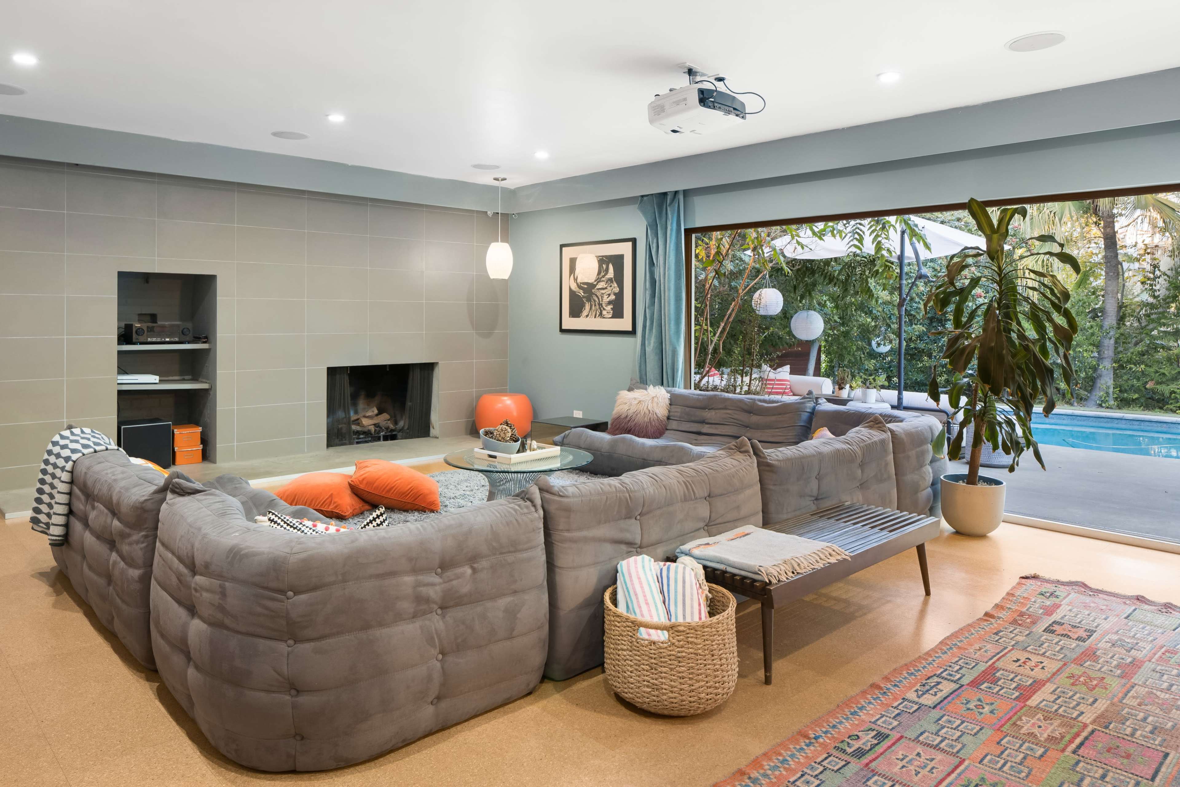 A modern living room features a gray sectional sofa, a fireplace, and large windows overlooking a pool area.