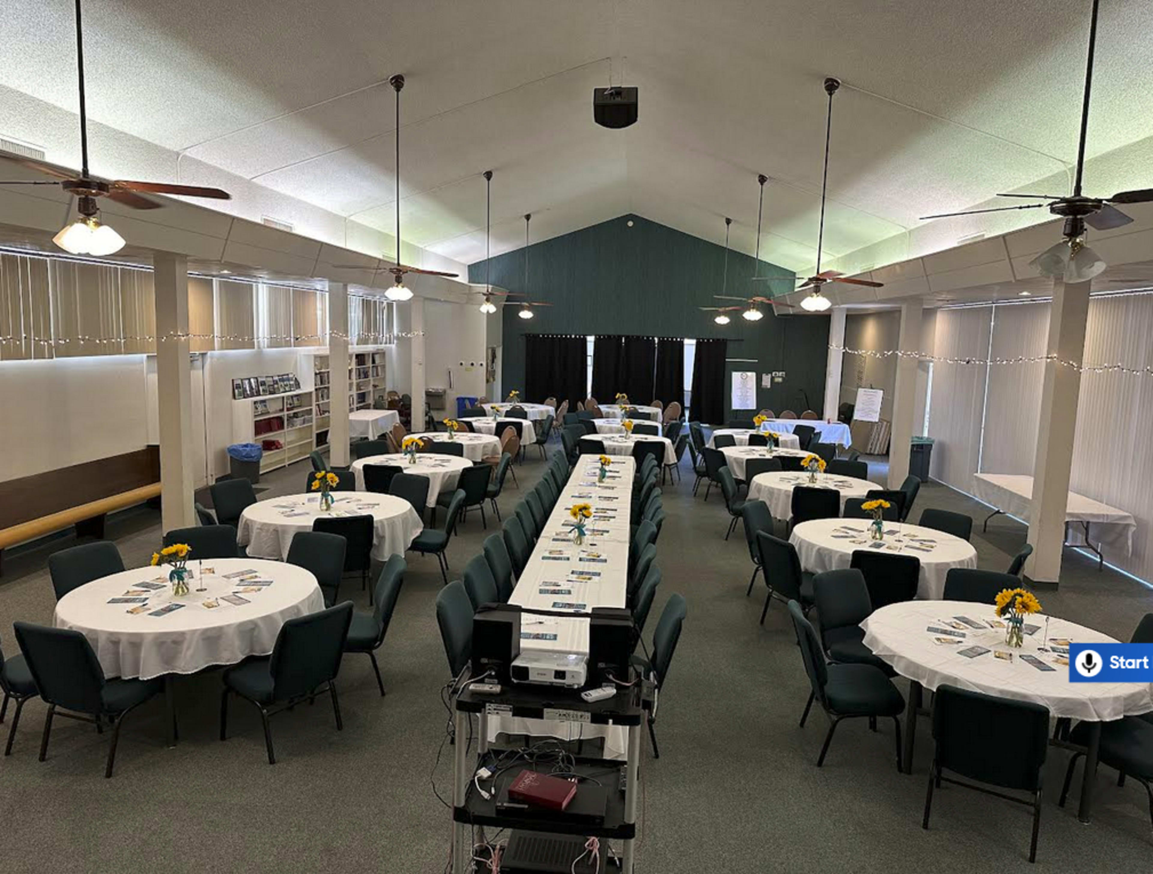 The image shows a spacious room set up for an event, featuring multiple round tables with white tablecloths and centerpieces of flowers, arranged in rows along a central aisle.