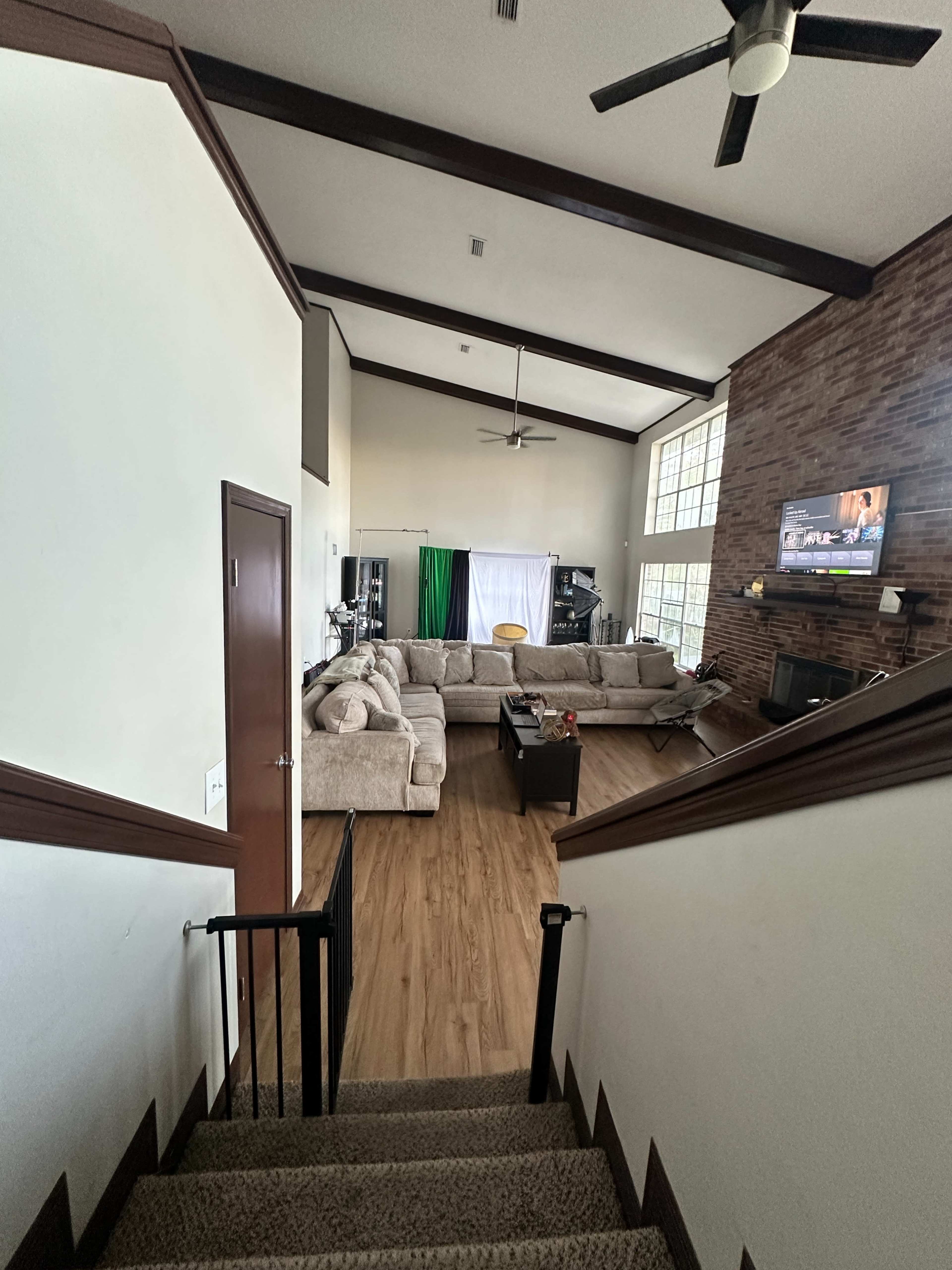 The image shows a staircase leading down to a spacious living room with a large sectional sofa, a coffee table, and a television against a brick wall.