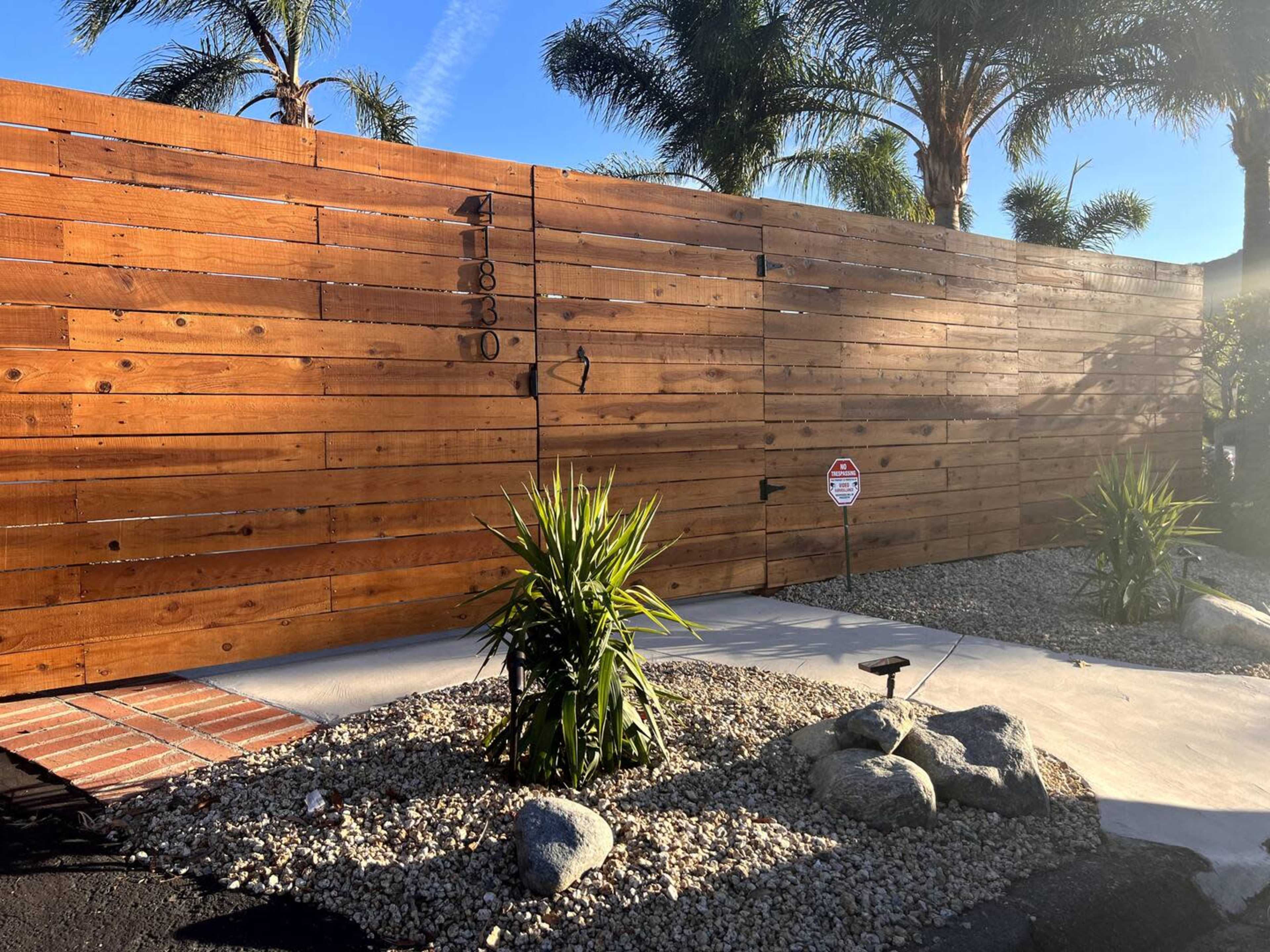 A wooden slat fence gates off a landscaped entrance with palm trees and ornamental rocks.