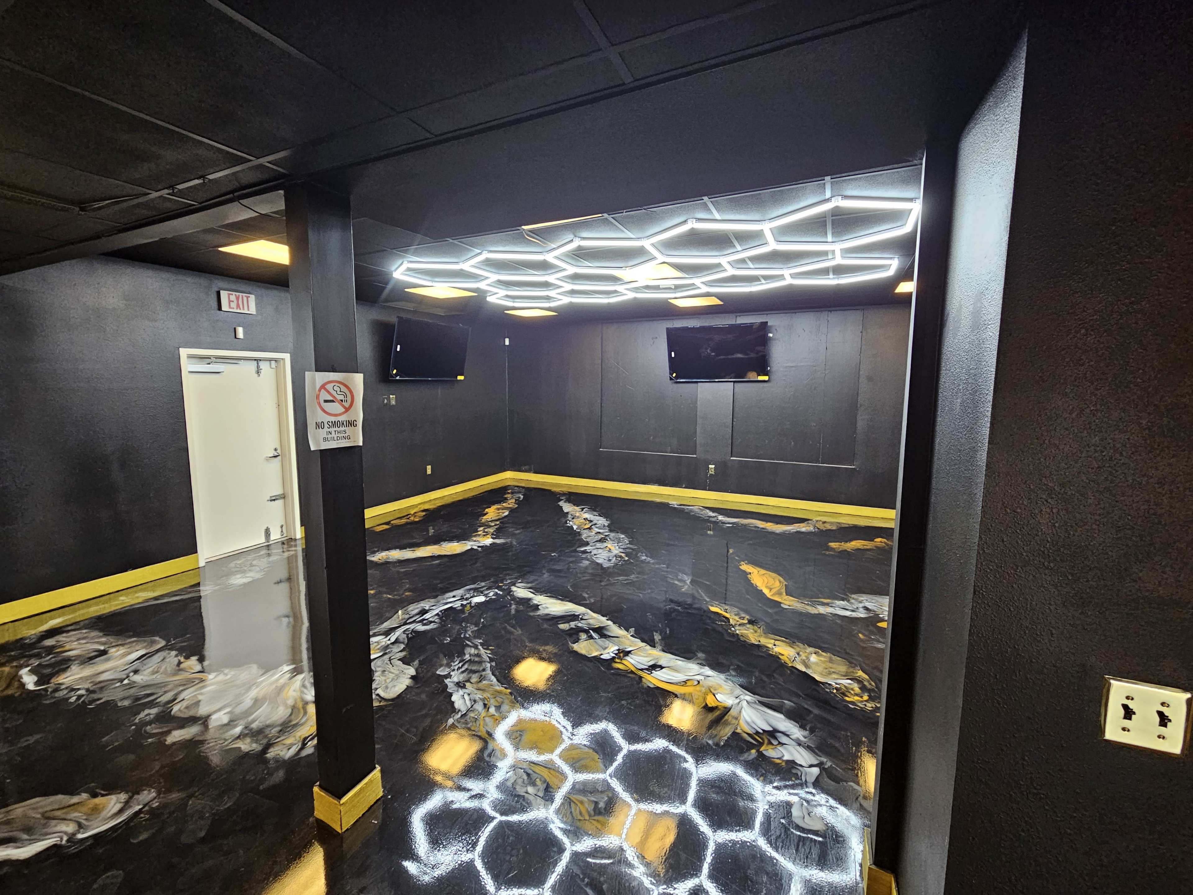 The image shows a modern room with black walls, a hexagonal-patterned illuminated ceiling, two mounted televisions, and glossy black-and-gold flooring.