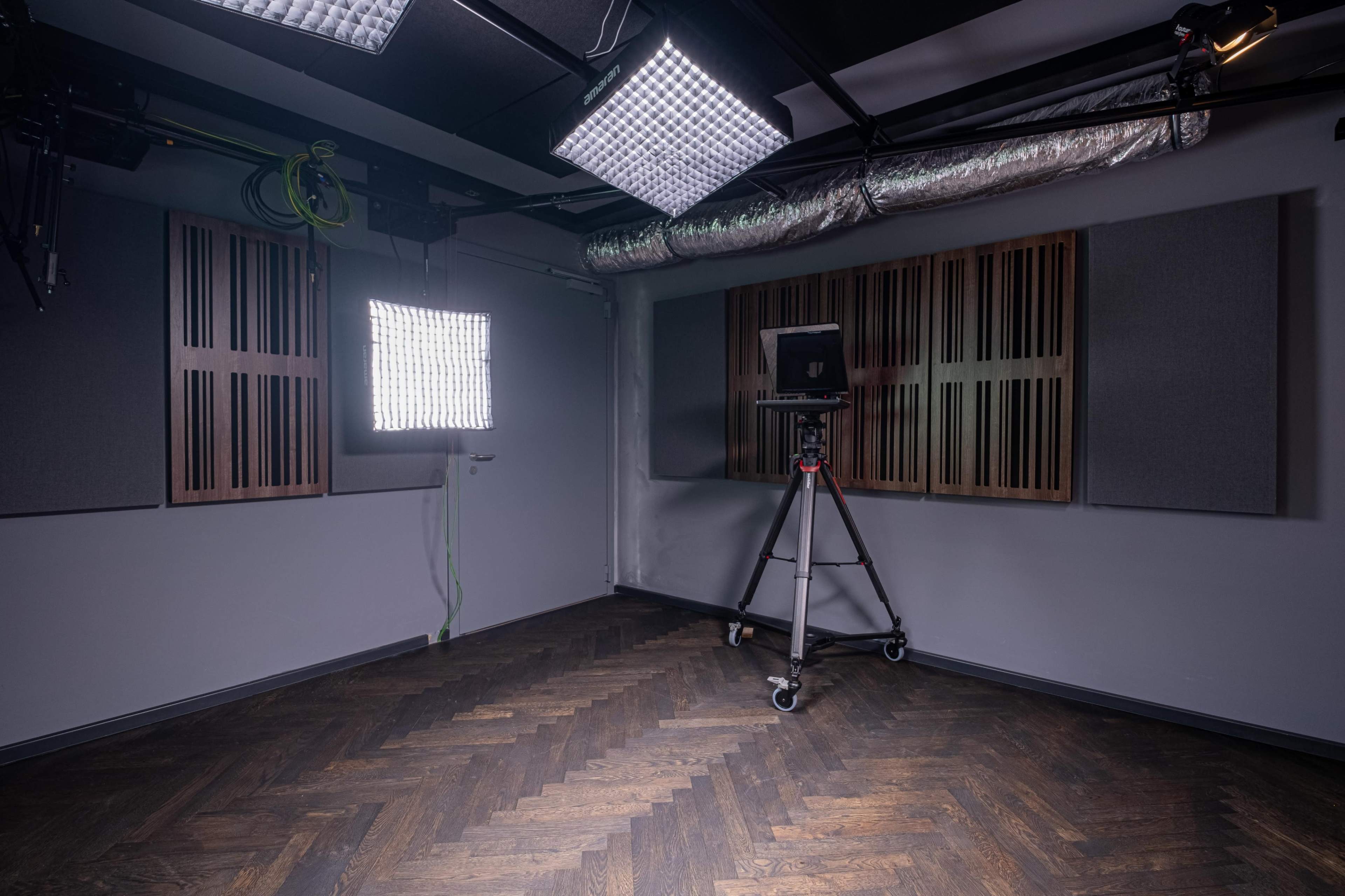 The image shows a studio room with acoustic panels on the walls, a tripod mounted camera, and overhead lighting.