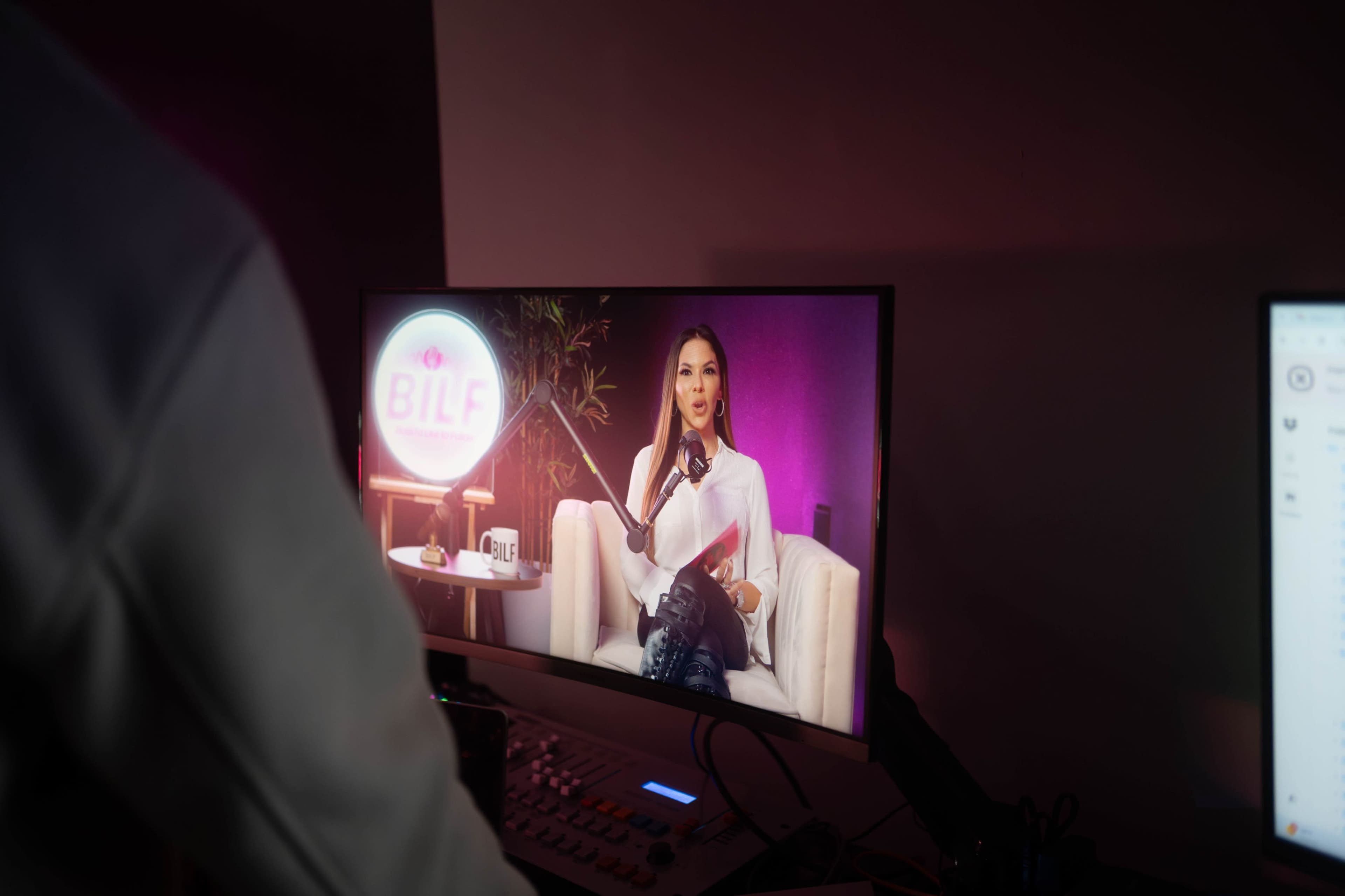 A woman sits on a sofa in front of a camera, speaking into a microphone while a neon sign with the acronym "BILF" glows in the background.