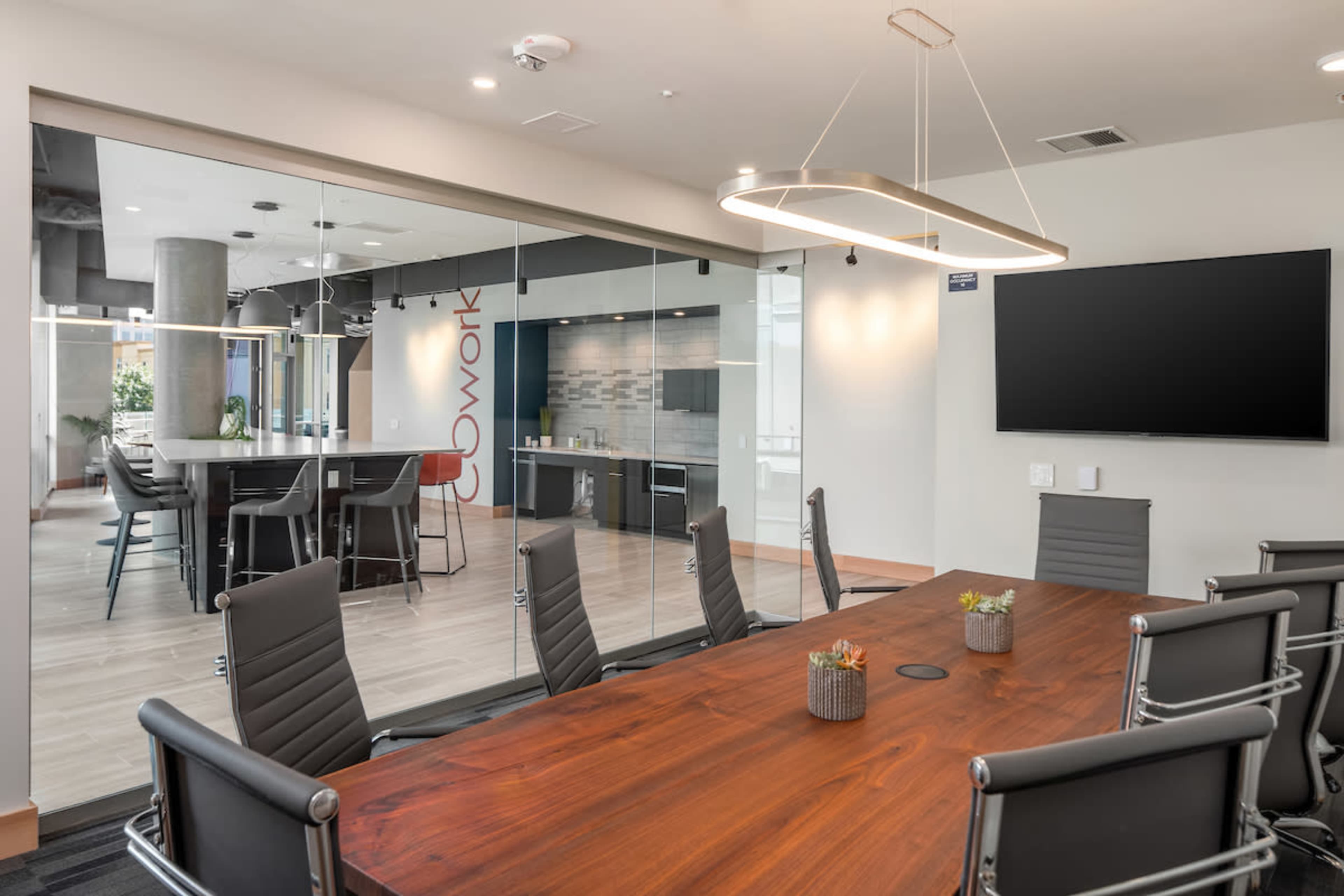 A modern conference room features a large wooden table surrounded by ergonomic chairs, with a wall-mounted screen and glass partitions leading to an adjacent workspace.