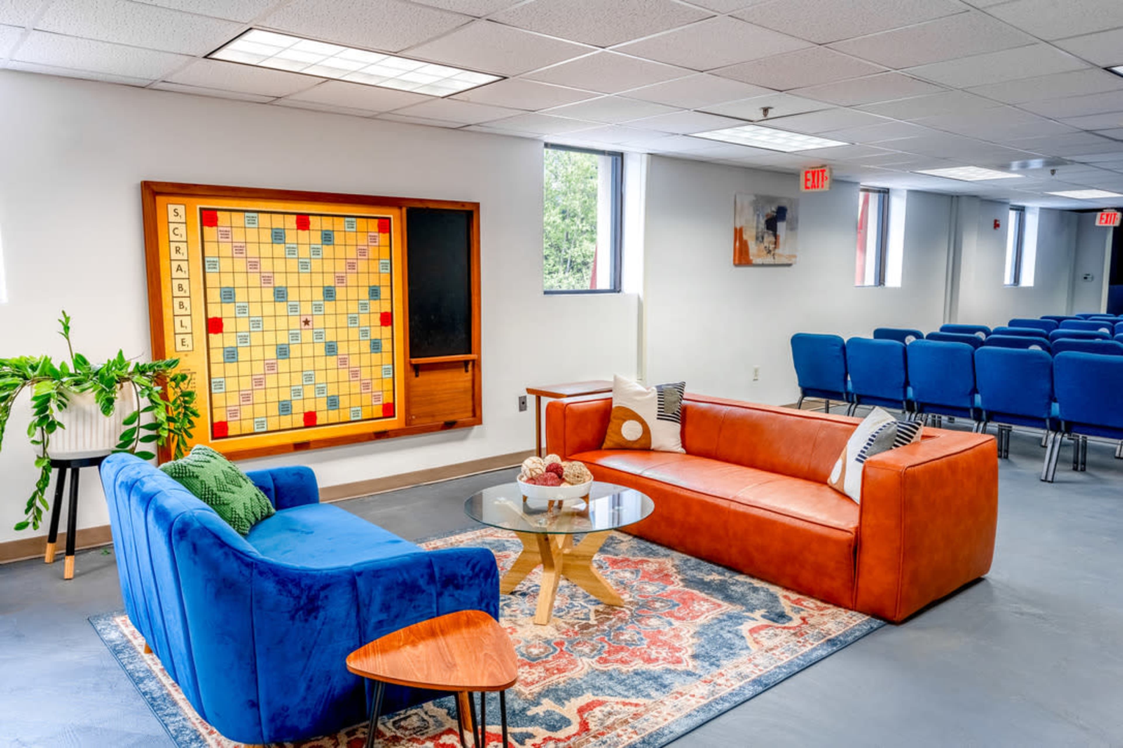 The image shows a modern room with blue and orange couches, a decorative rug, a coffee table with snacks, a large Scrabble board on the wall, and rows of blue chairs arranged in the background.