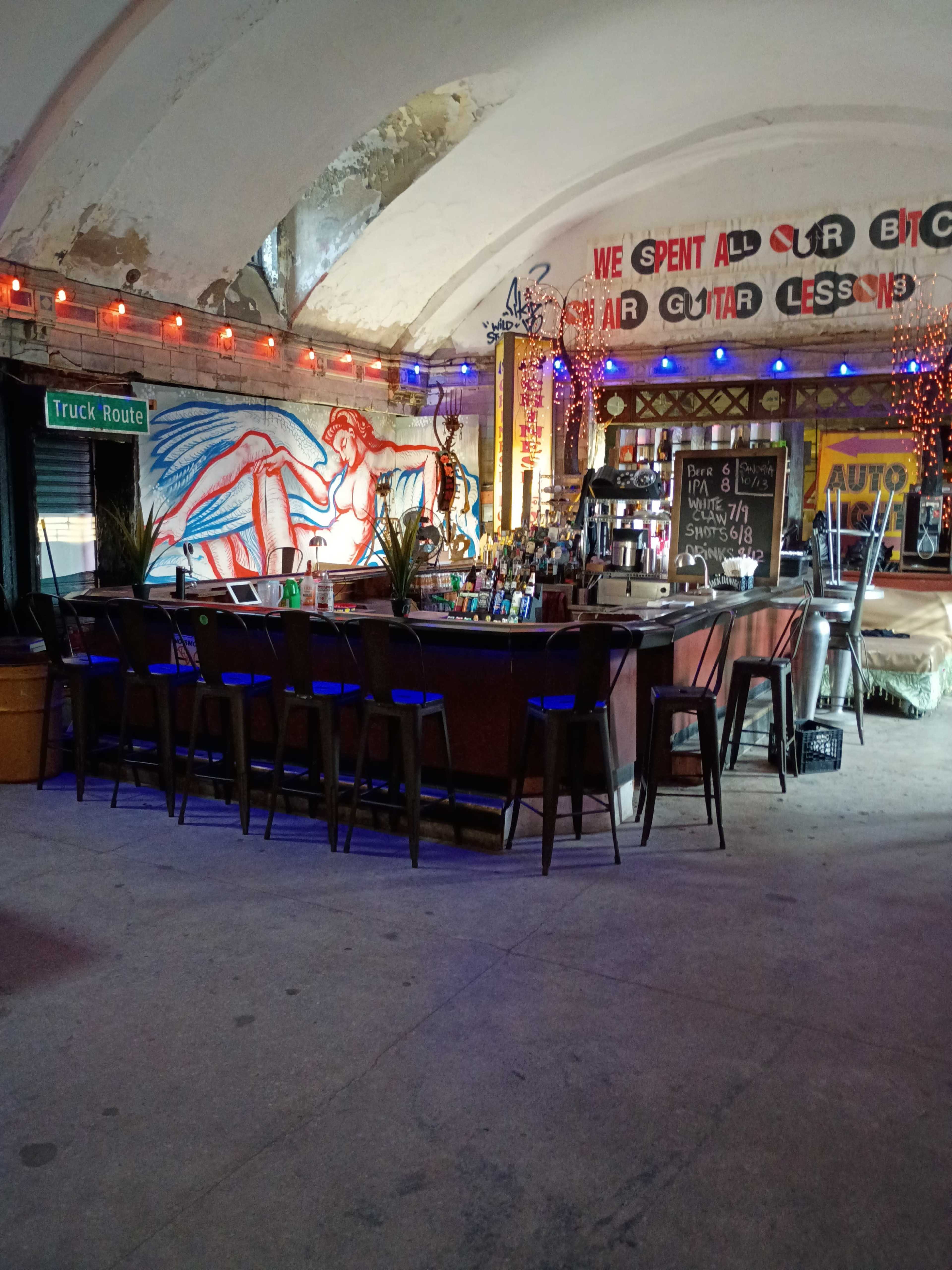 The image shows the interior of a bar featuring a large mural and colorful lighting with a long counter and several bar stools.