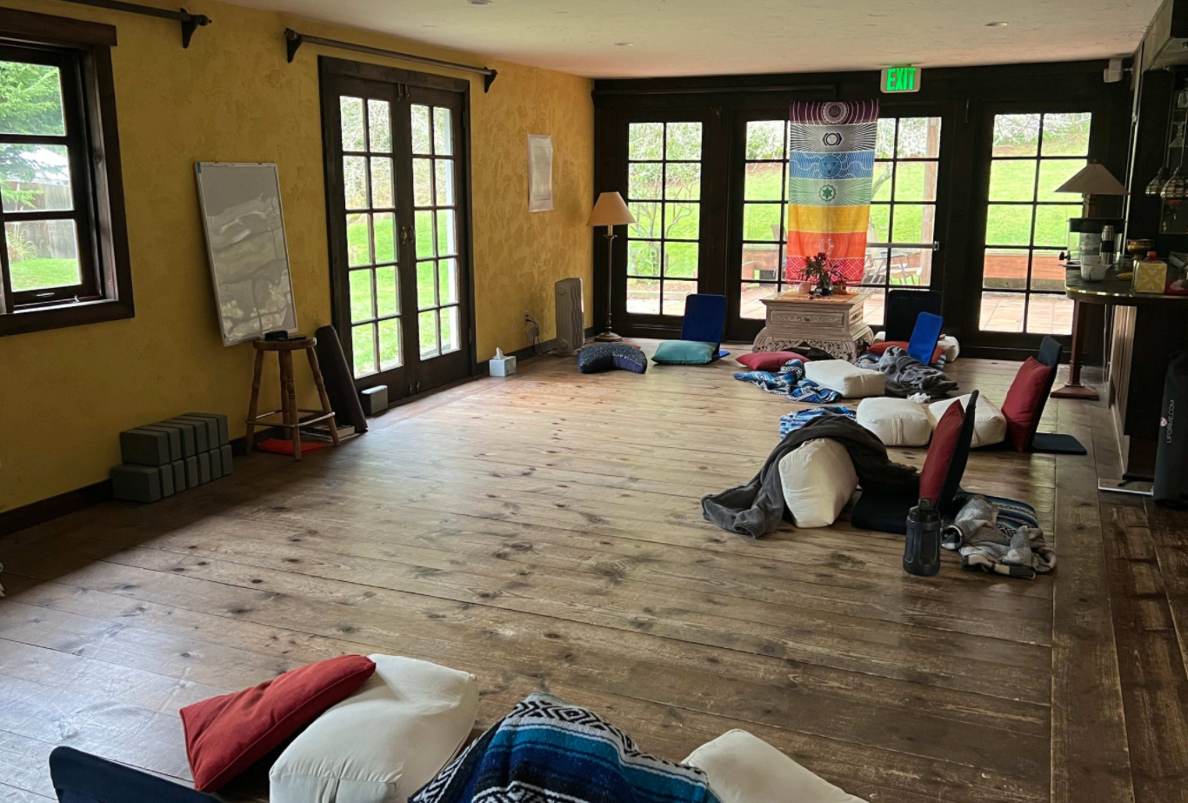 A spacious room with wooden floors, arranged with cushions and pillows, and large windows leading to an outdoor area.