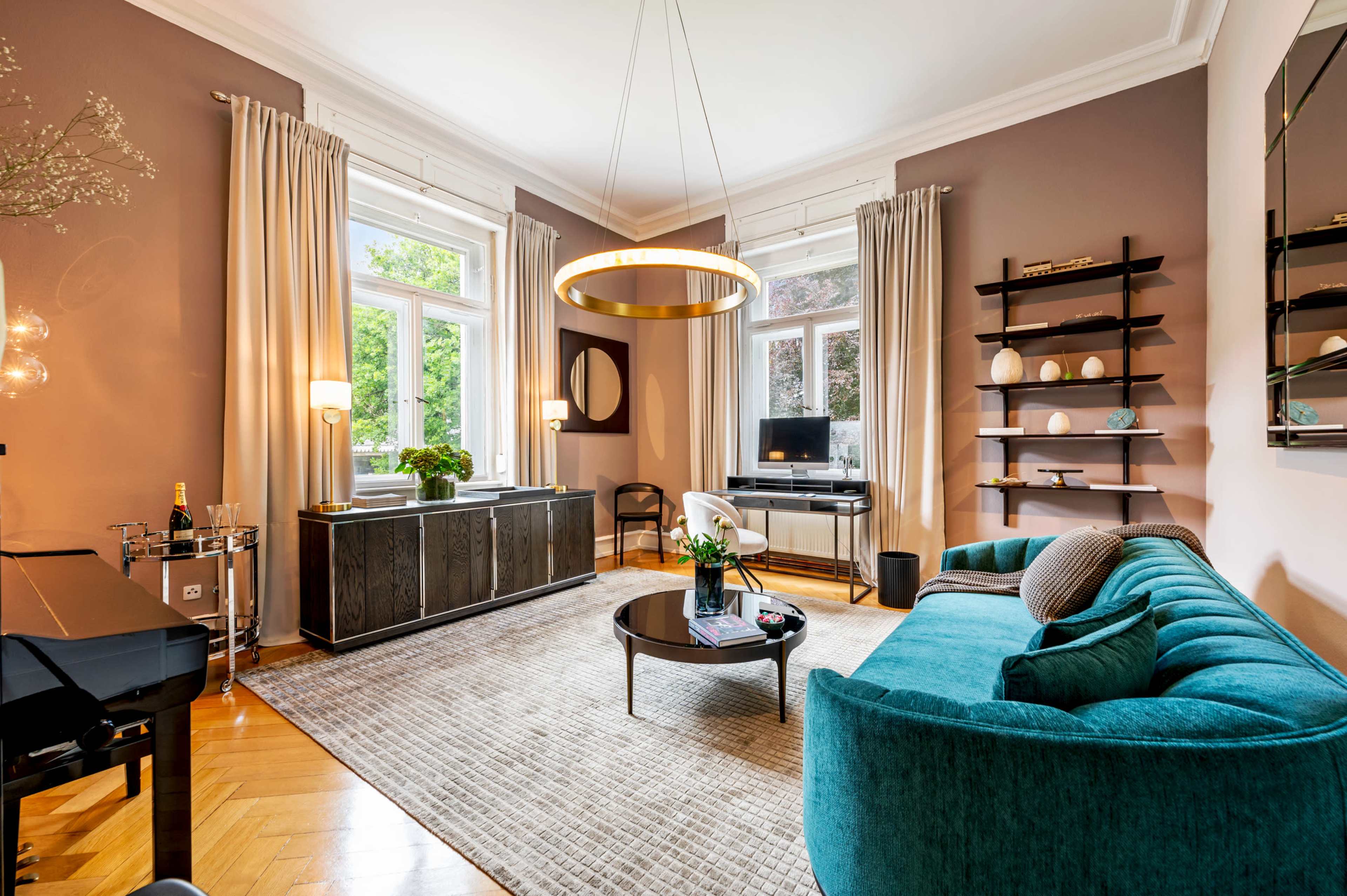 The image shows a cozy living room featuring a teal sofa, large windows with drapes, a round coffee table, and a combination of modern furniture and decorative items.