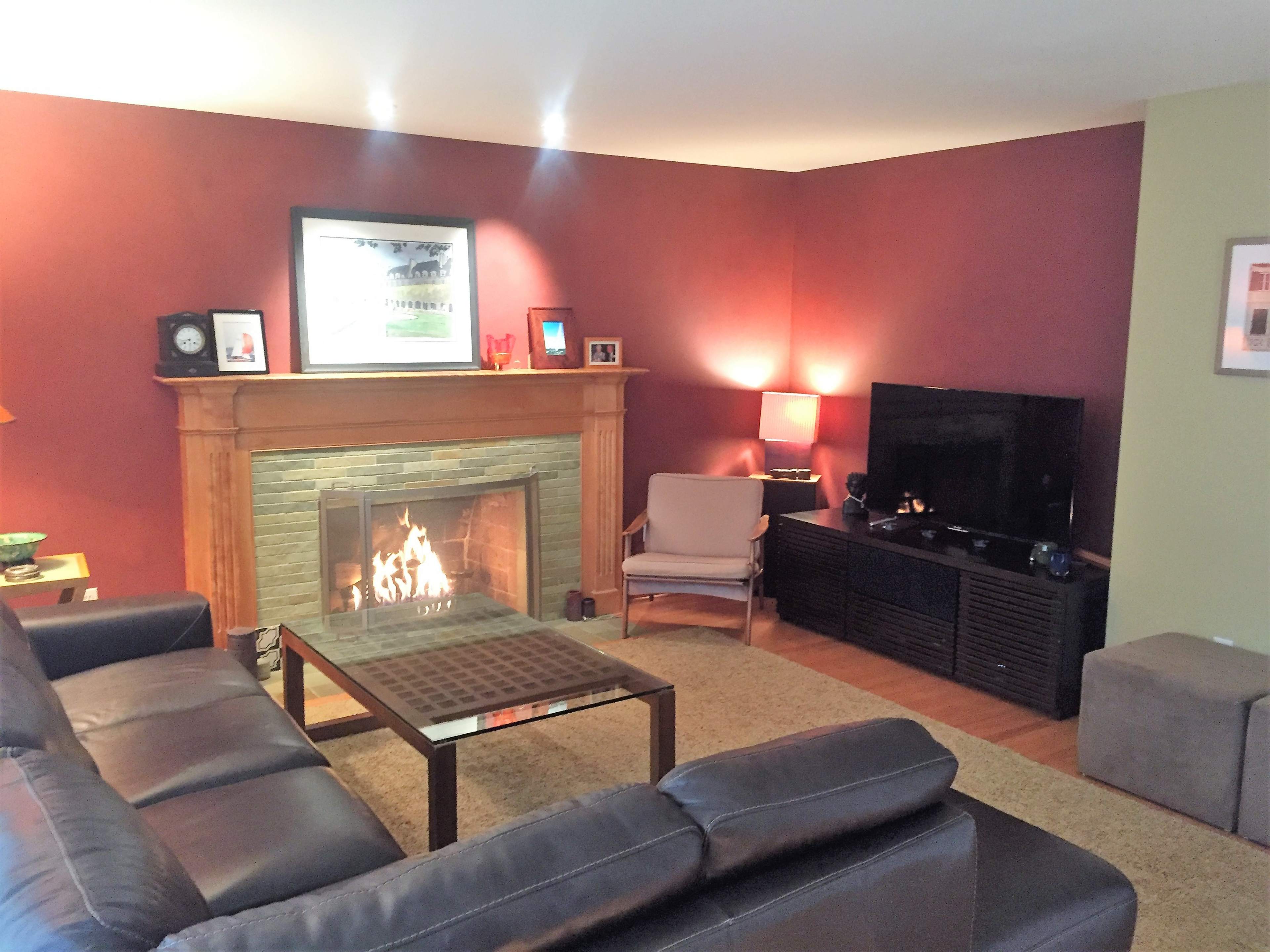 The living room features a stone fireplace with a lit fire, a large leather sectional sofa, and a television mounted on a wall across from the seating area.