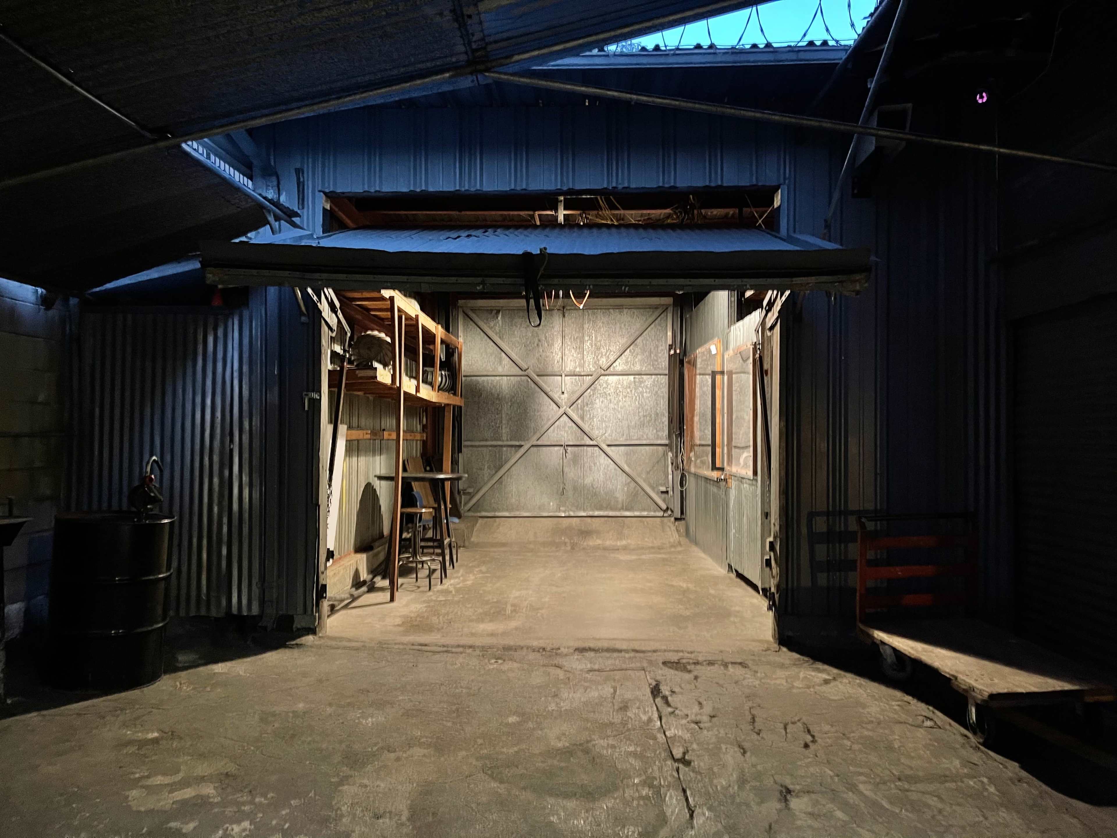 A dimly lit industrial space with metal walls, a large garage door, and wooden shelving units along one side.