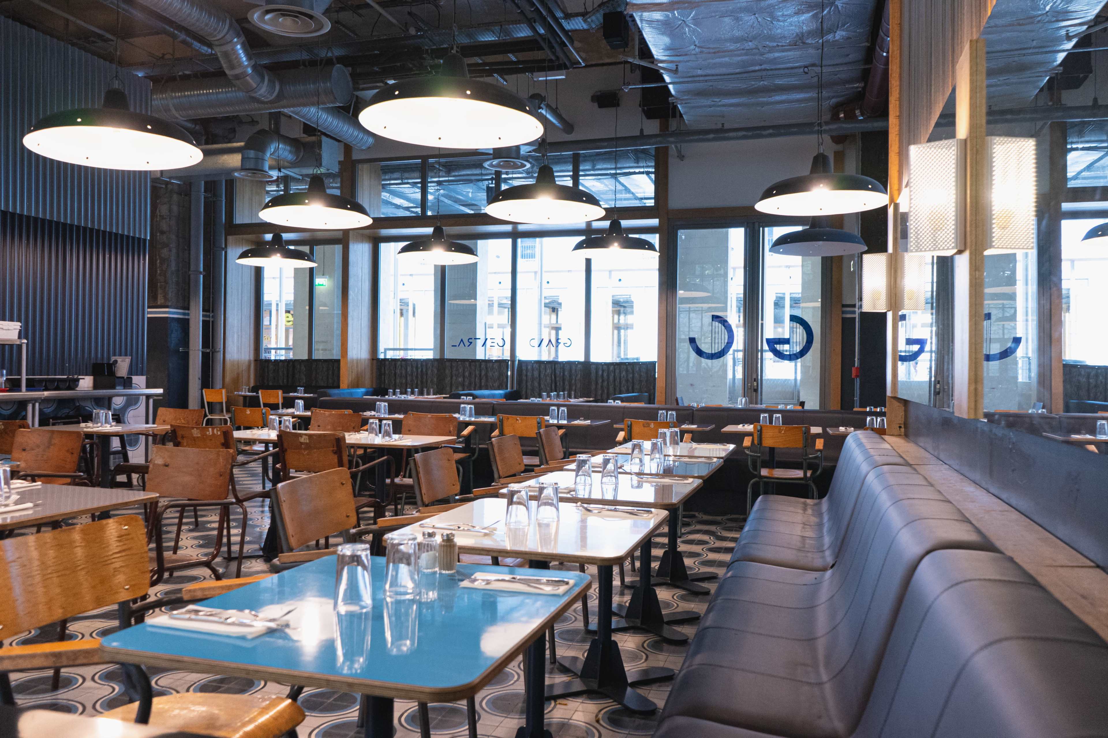 The interior of a modern restaurant features wooden tables, leather benches, and large windows letting in natural light.