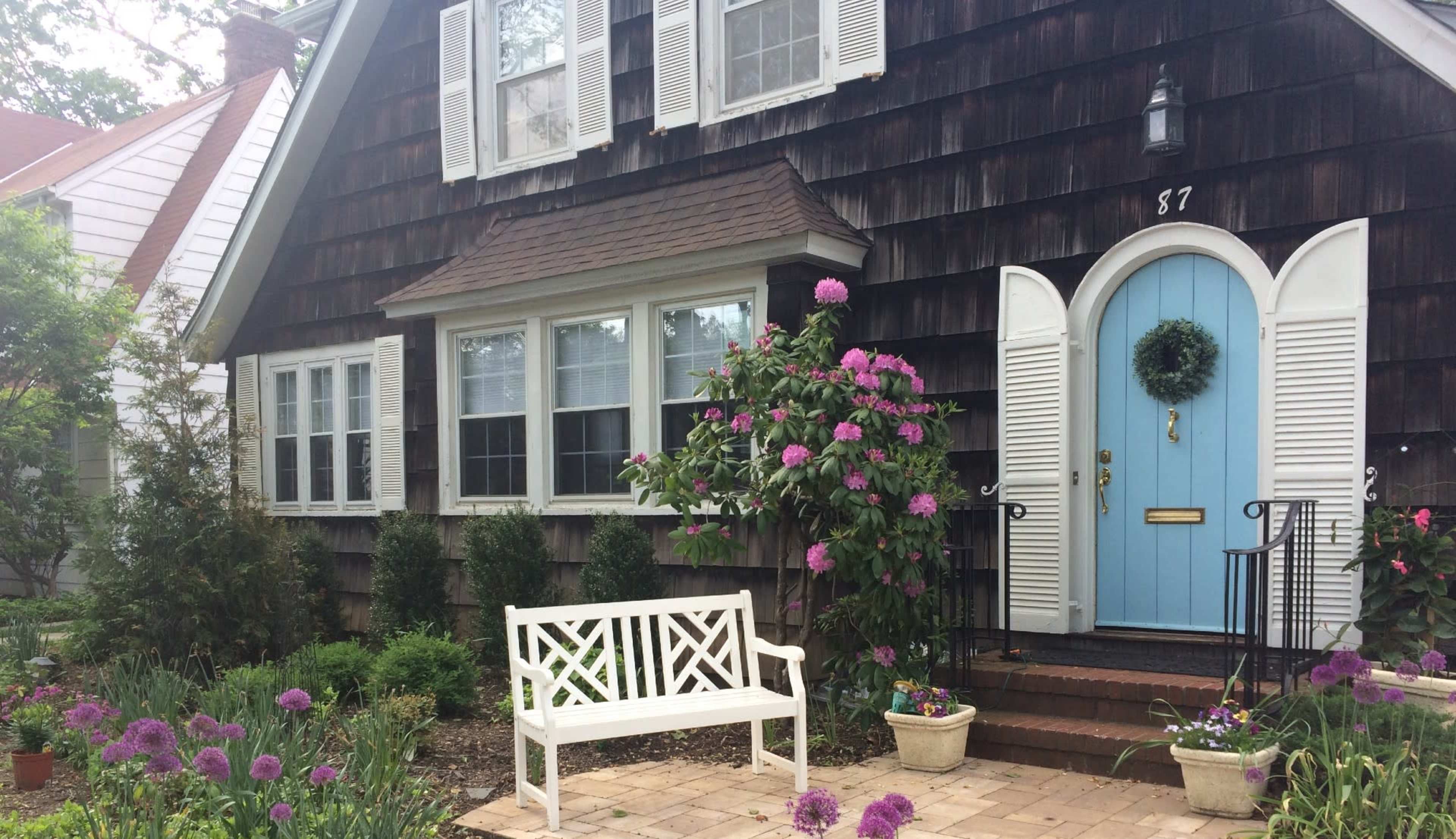 A charming house with a blue front door and flower beds filled with purple blooms in front of a wooden-shingled exterior.