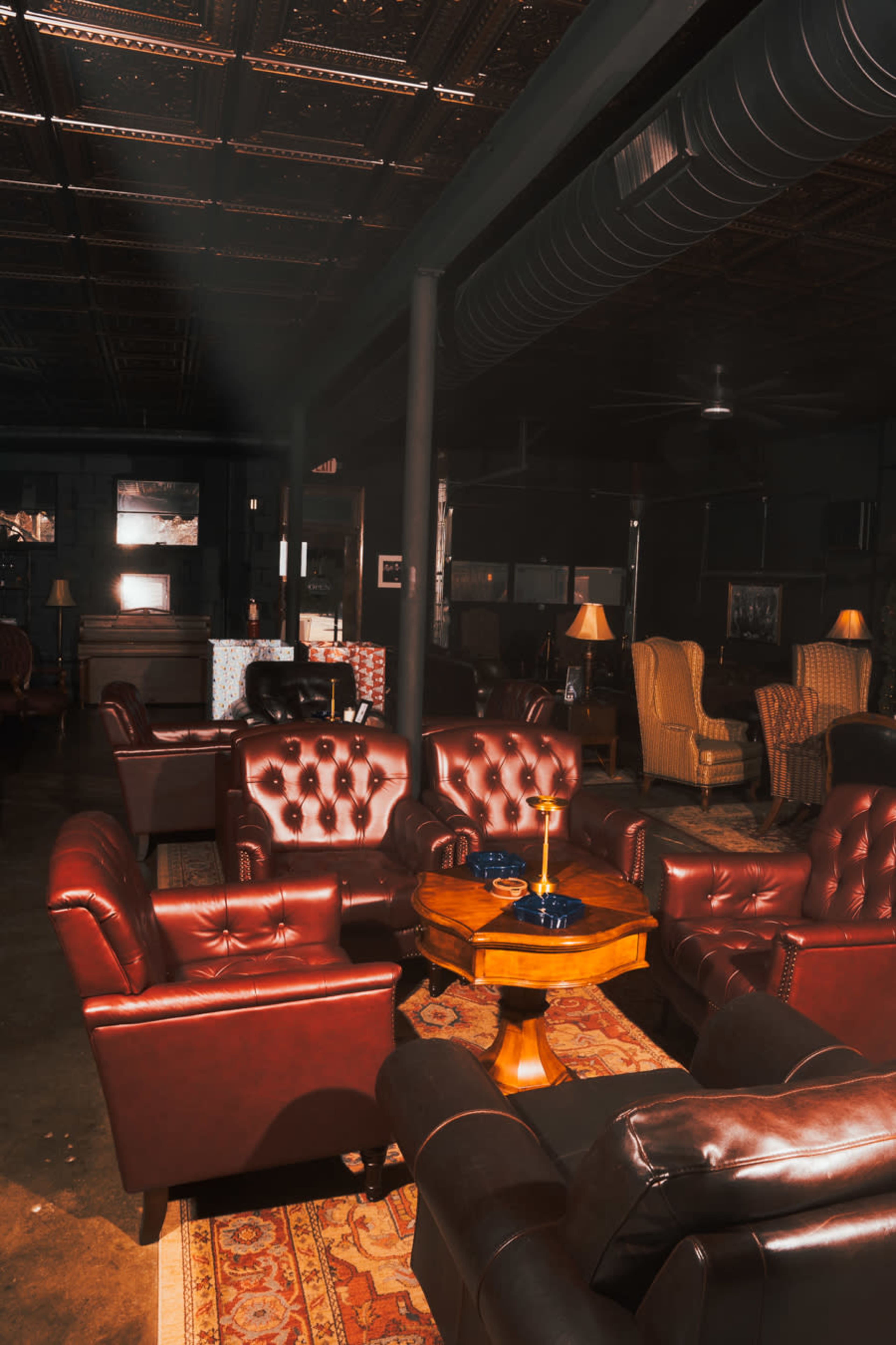 The image shows a dimly lit lounge area with dark leather chairs arranged around a wooden table, set on a patterned rug.
