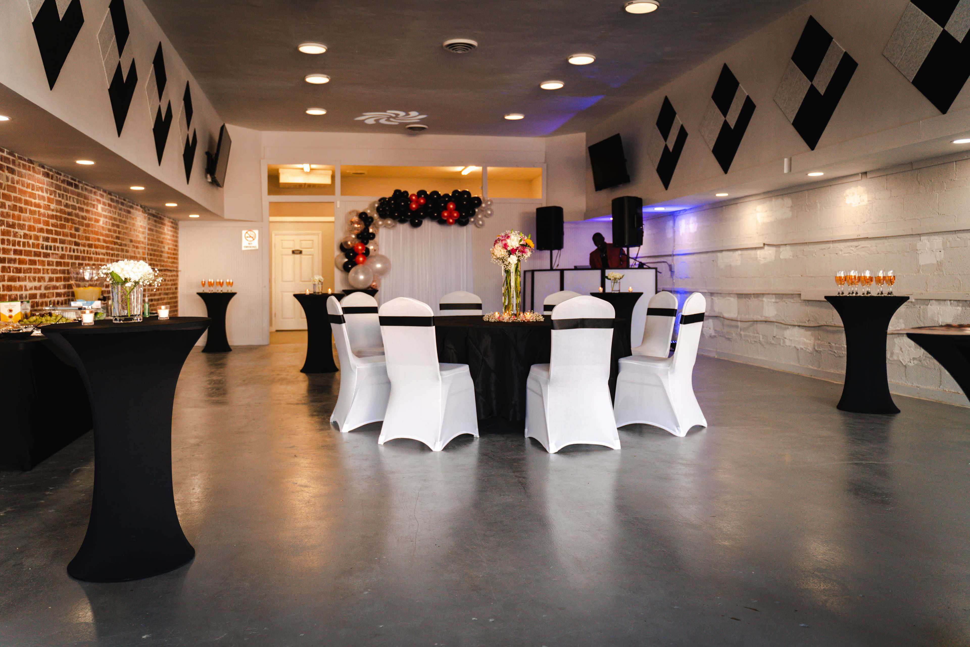 A spacious event room features black and white tablecloths arranged with chairs, decorated with floral centerpieces and a backdrop of balloons.
