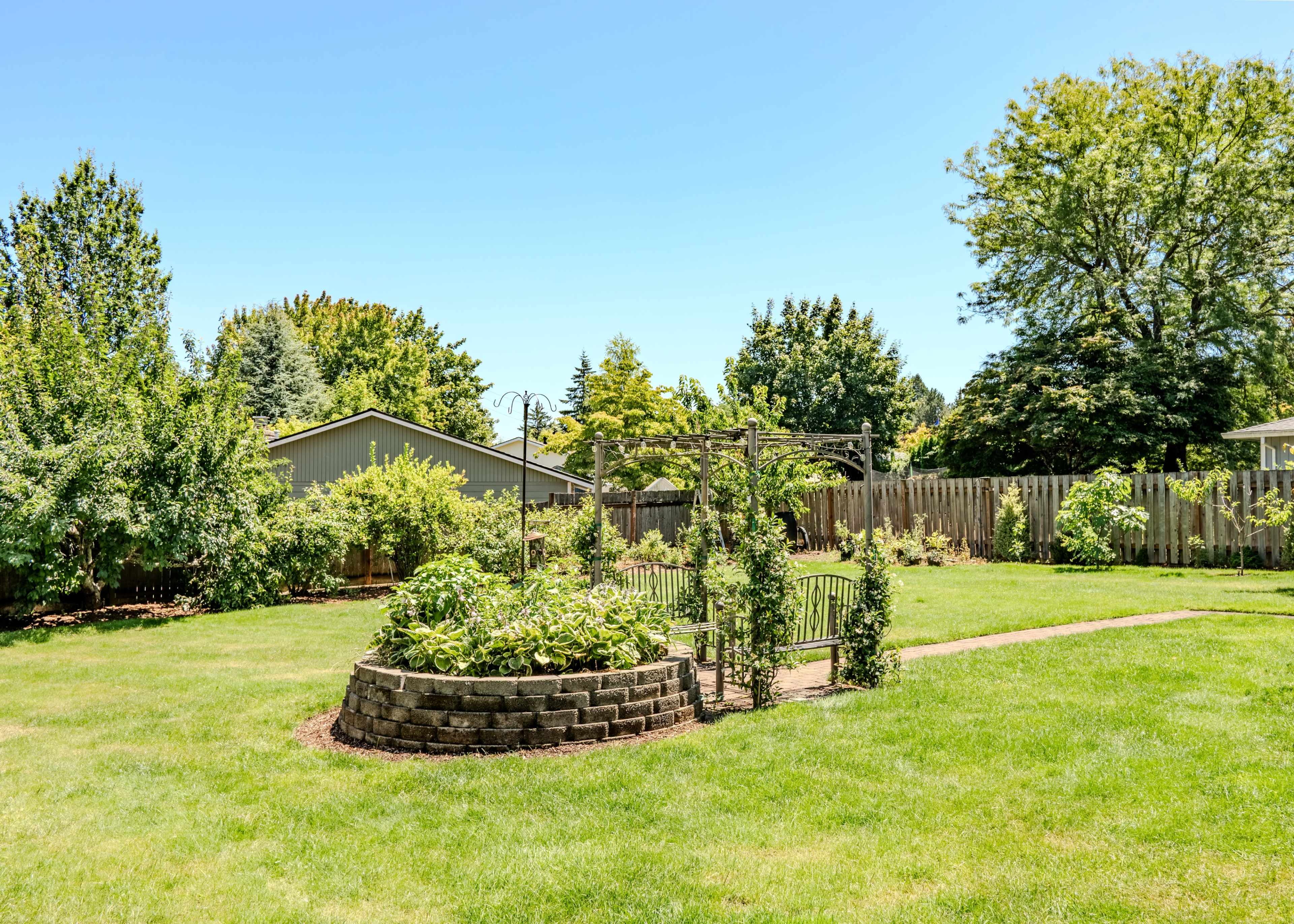 The image shows a spacious backyard with a circular garden surrounded by greenery and a wooden fence in the background.