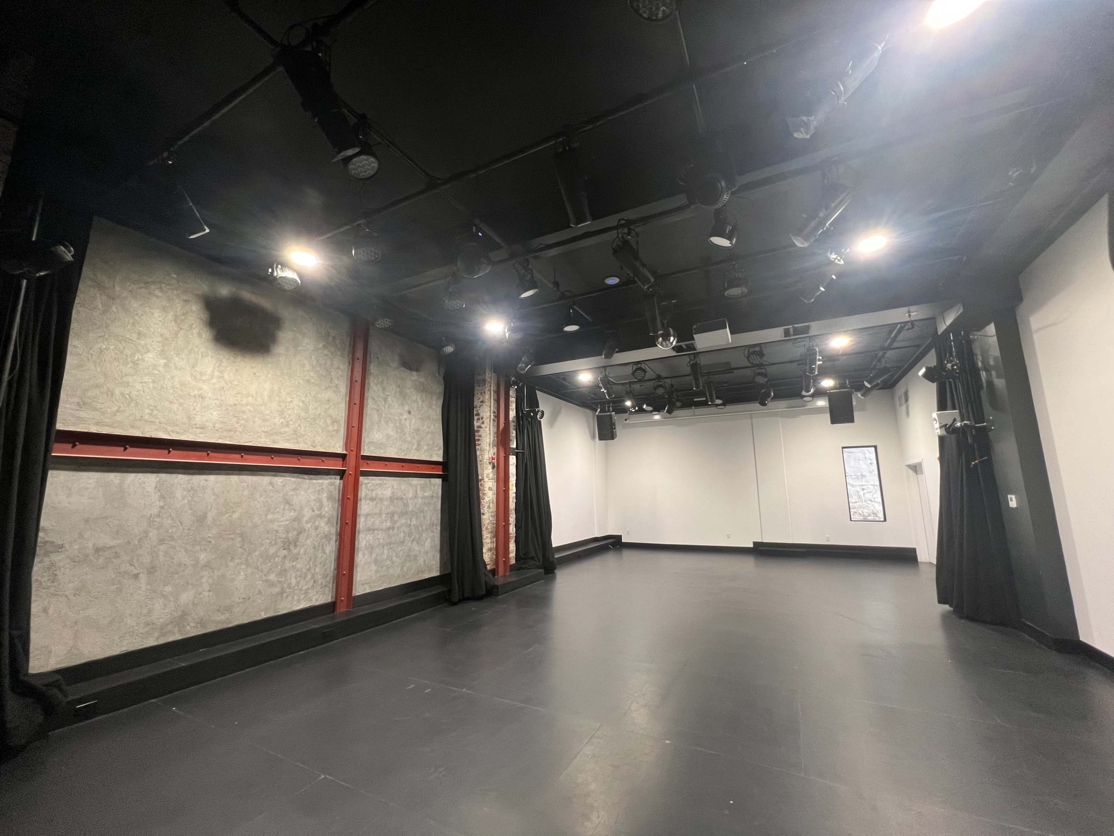 The image shows a spacious, well-lit black box theater with a bare stage, textured walls, and adjustable lighting fixtures.