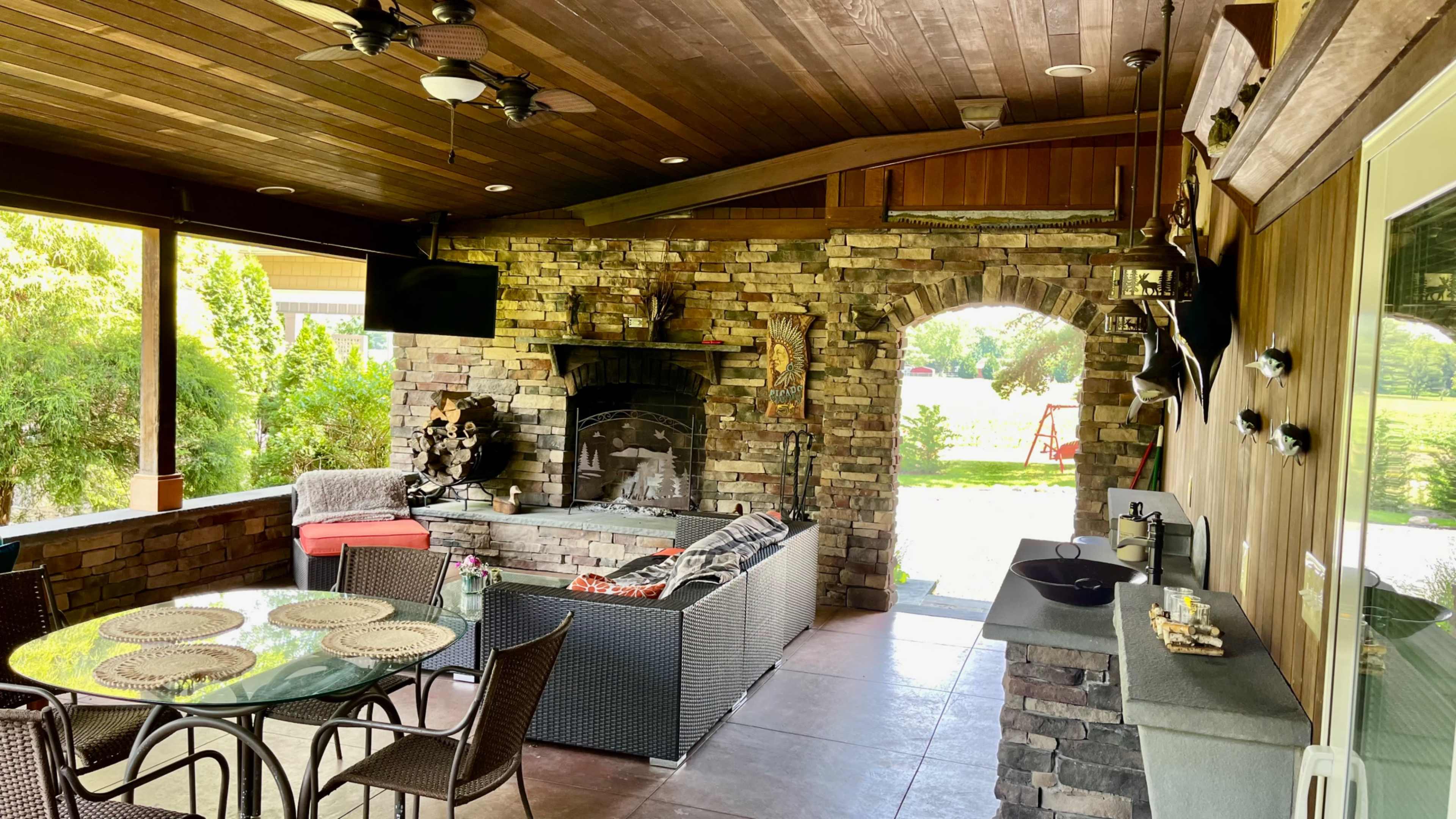 The image shows a covered outdoor patio with stone walls, a fireplace, a dining table, and comfortable seating arrangements.