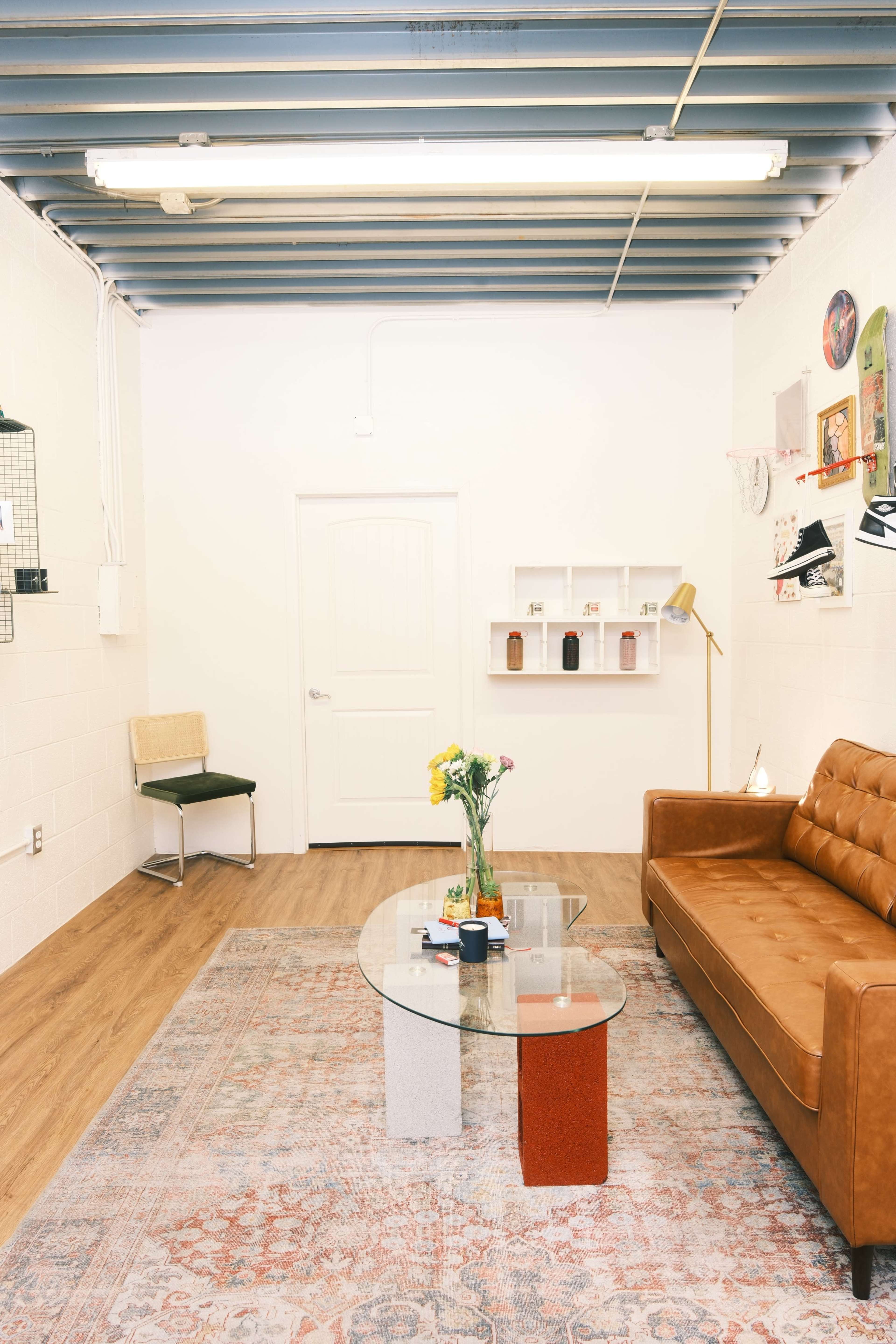The image shows a minimalist room with a brown leather sofa, a clear glass coffee table, and a small shelf on the wall, set against a backdrop of light-colored walls and wooden flooring.