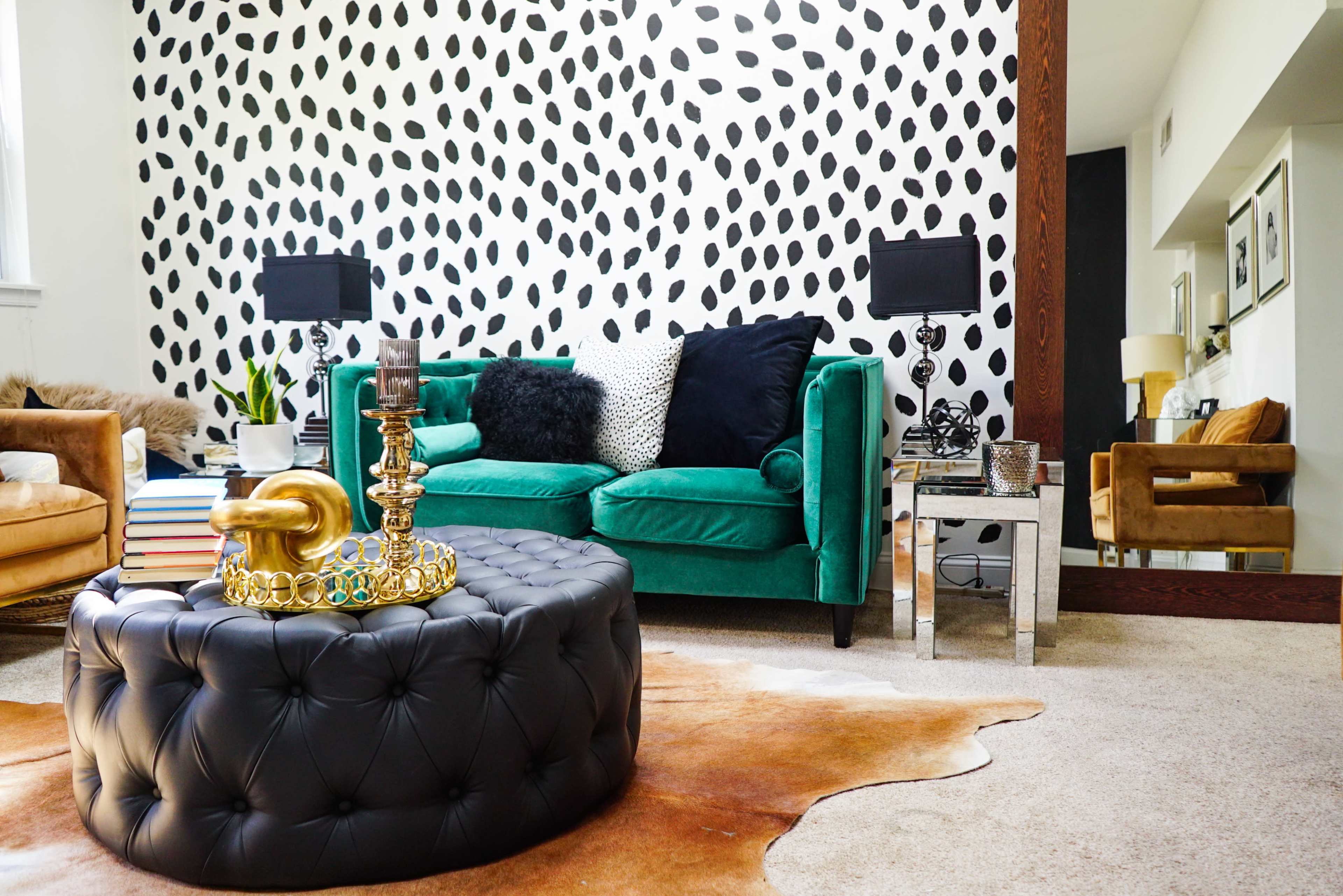 A modern living room with a green velvet sofa, a black tufted coffee table, a decorative gold candle holder, and a unique wallpaper design of black leaves against a white background.