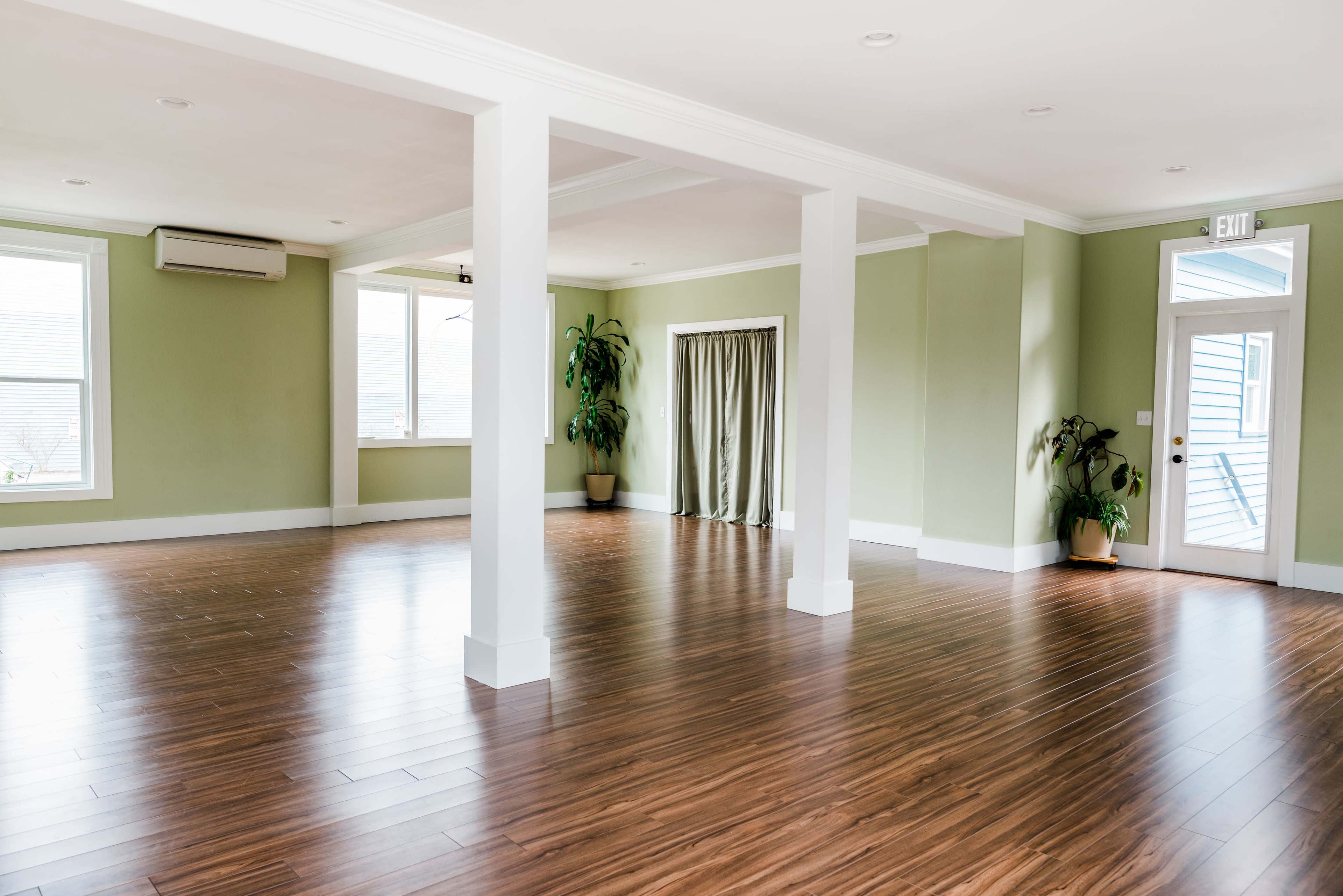 The image shows an empty room with wooden flooring, green walls, and several windows, featuring plants in the corners and a doorway leading outside.