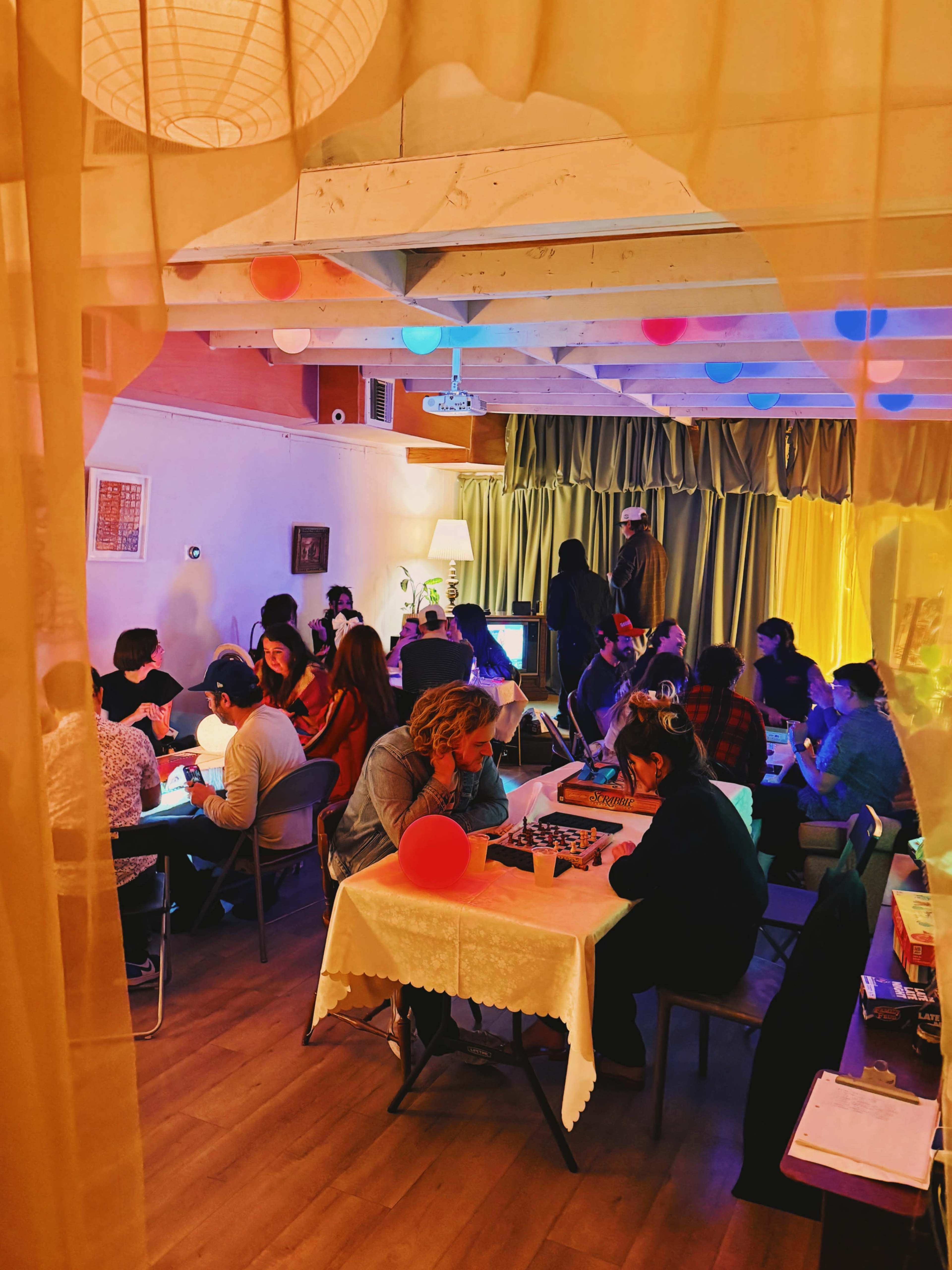 A group of people is gathered in a brightly lit room, engaged in various activities around tables, while colorful decorations hang from the ceiling.