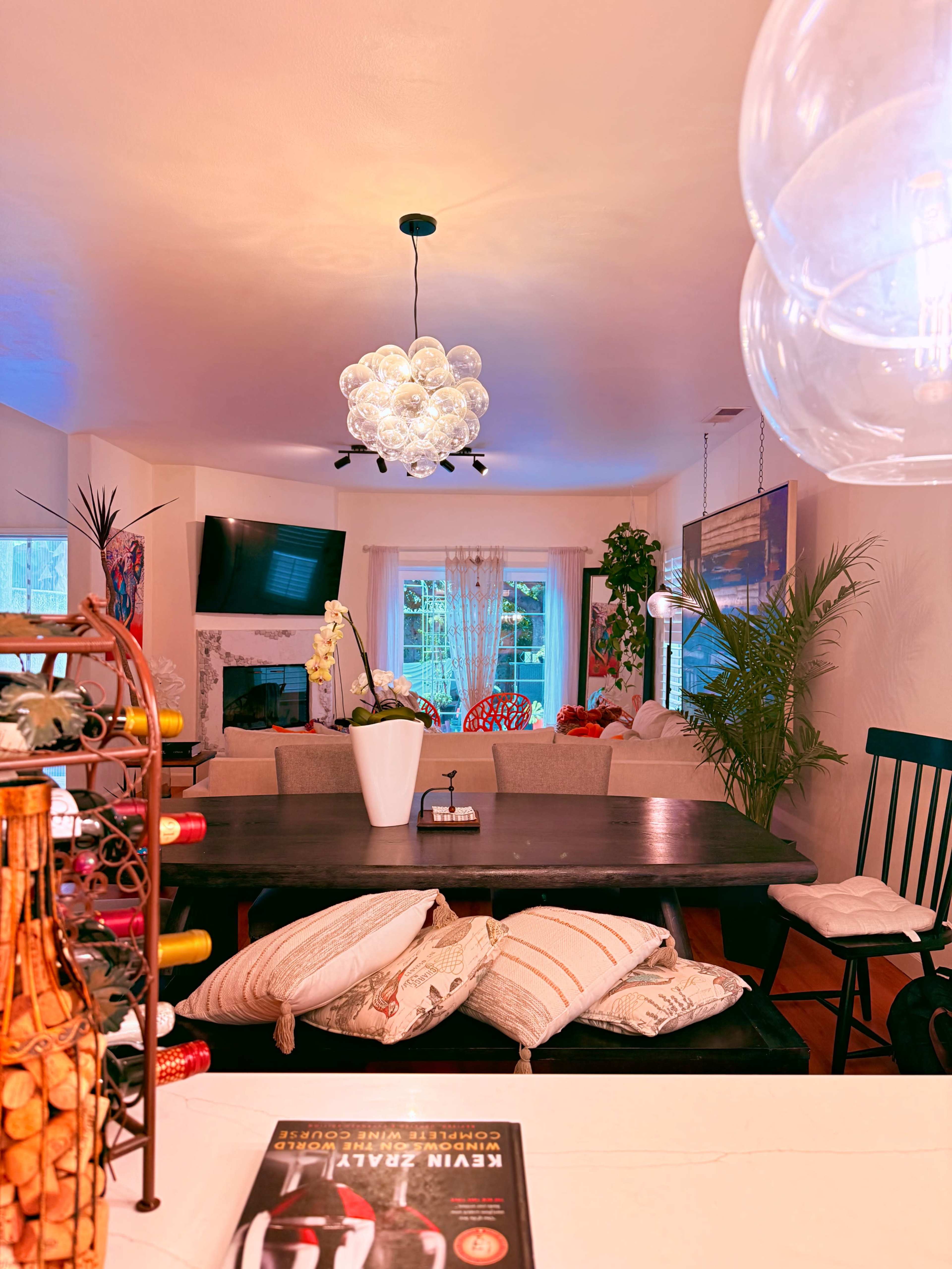The image shows a bright dining area with a dark wooden table, decorative bottles, and a modern light fixture, leading to a cozy living space in the background.