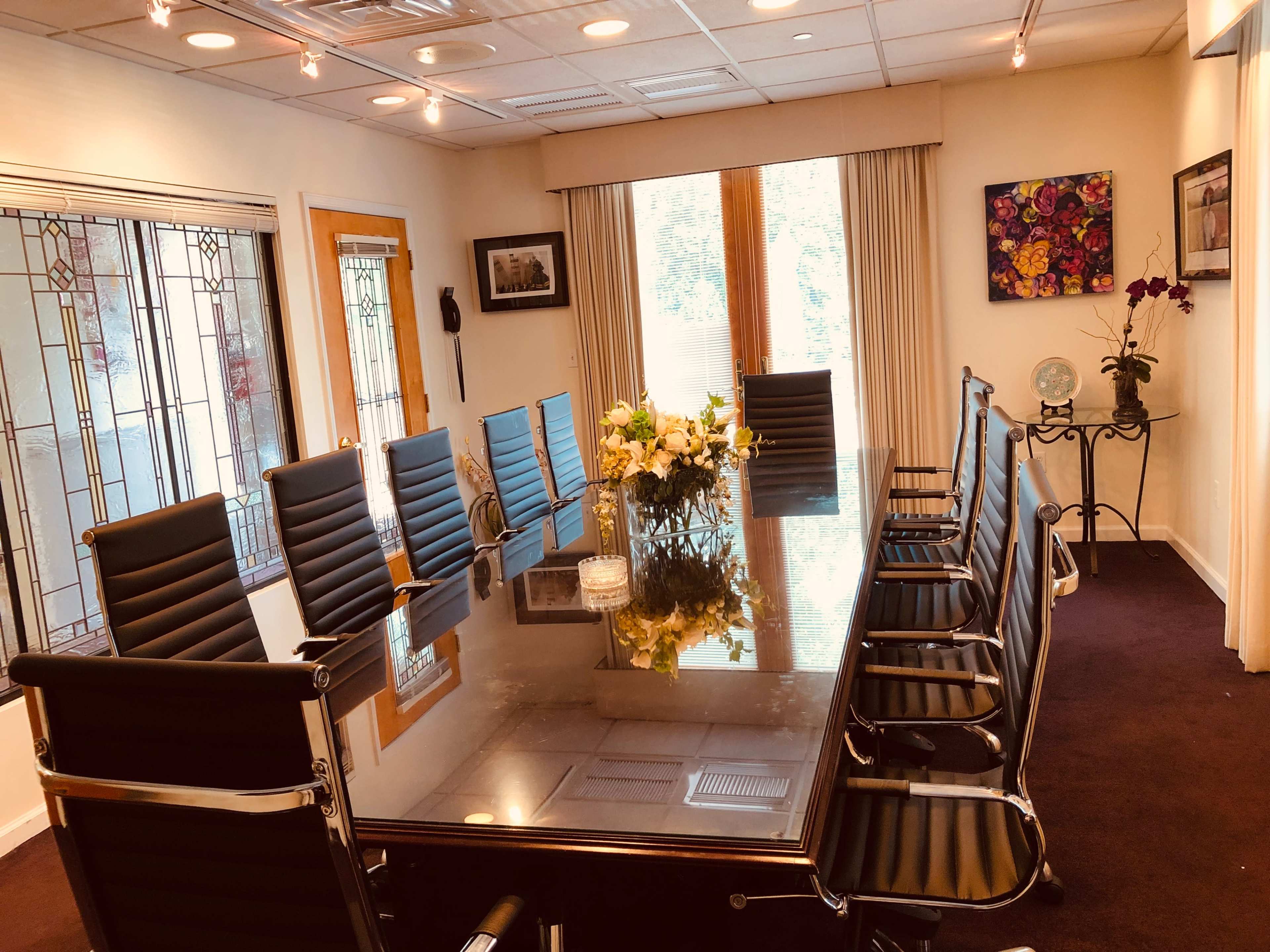 A conference room features a long glass table surrounded by black office chairs, with a floral centerpiece and large windows letting in natural light.