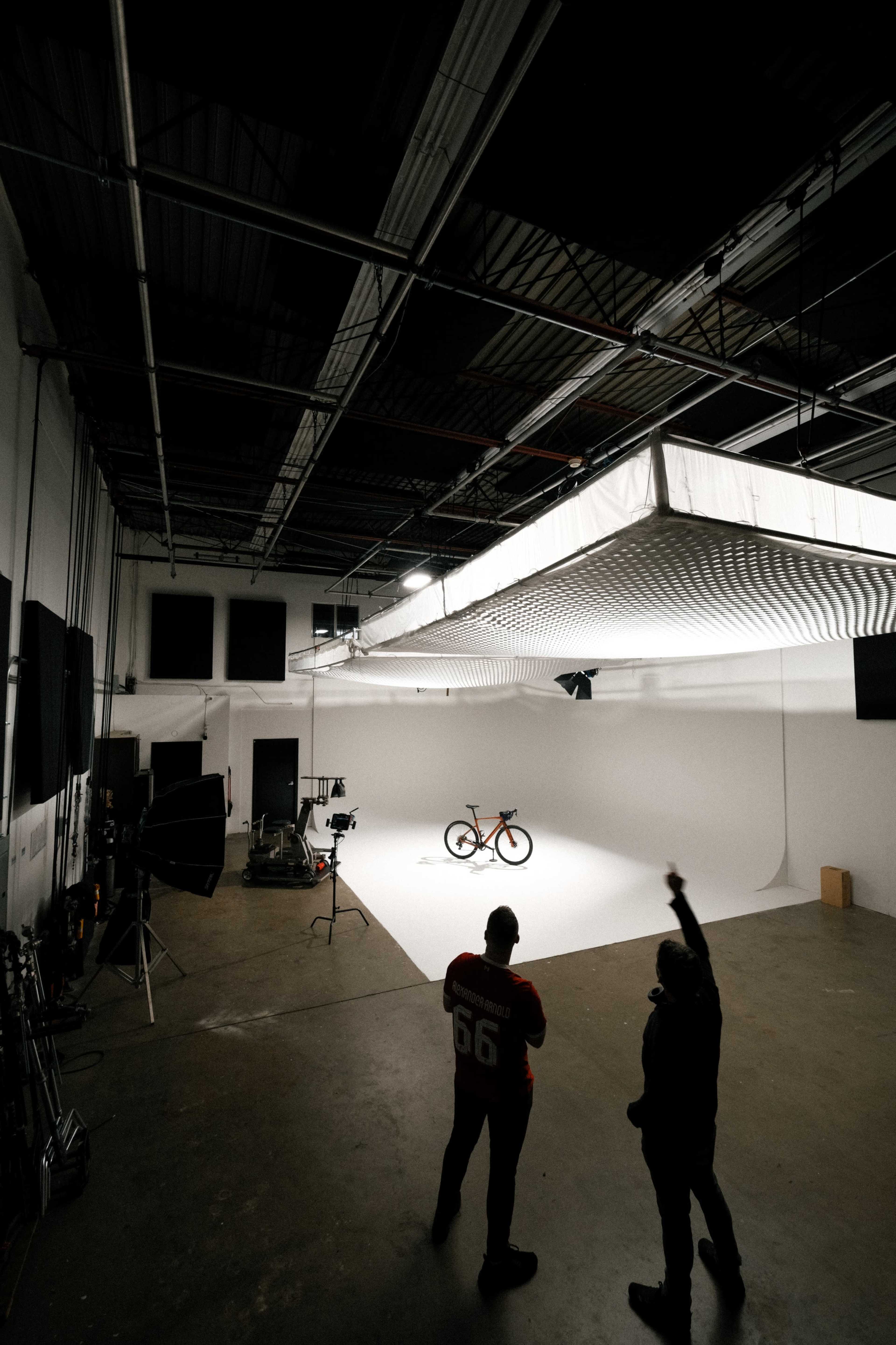 Two individuals stand in a photography studio, observing a bicycle positioned on a white backdrop under bright lights.