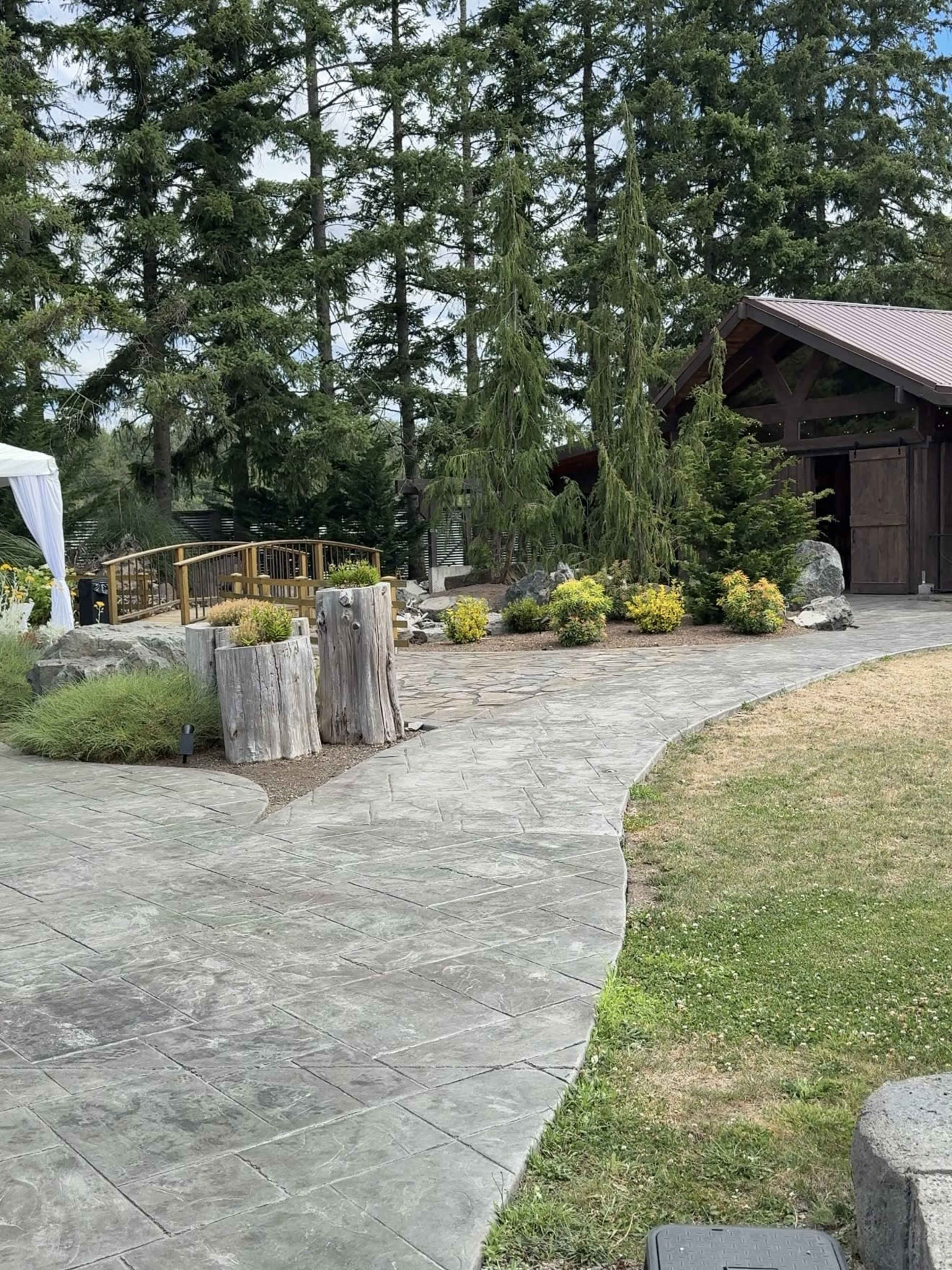 The image shows a landscaped outdoor area featuring a curved stone pathway, wooden accents, and greenery, with a rustic building in the background.