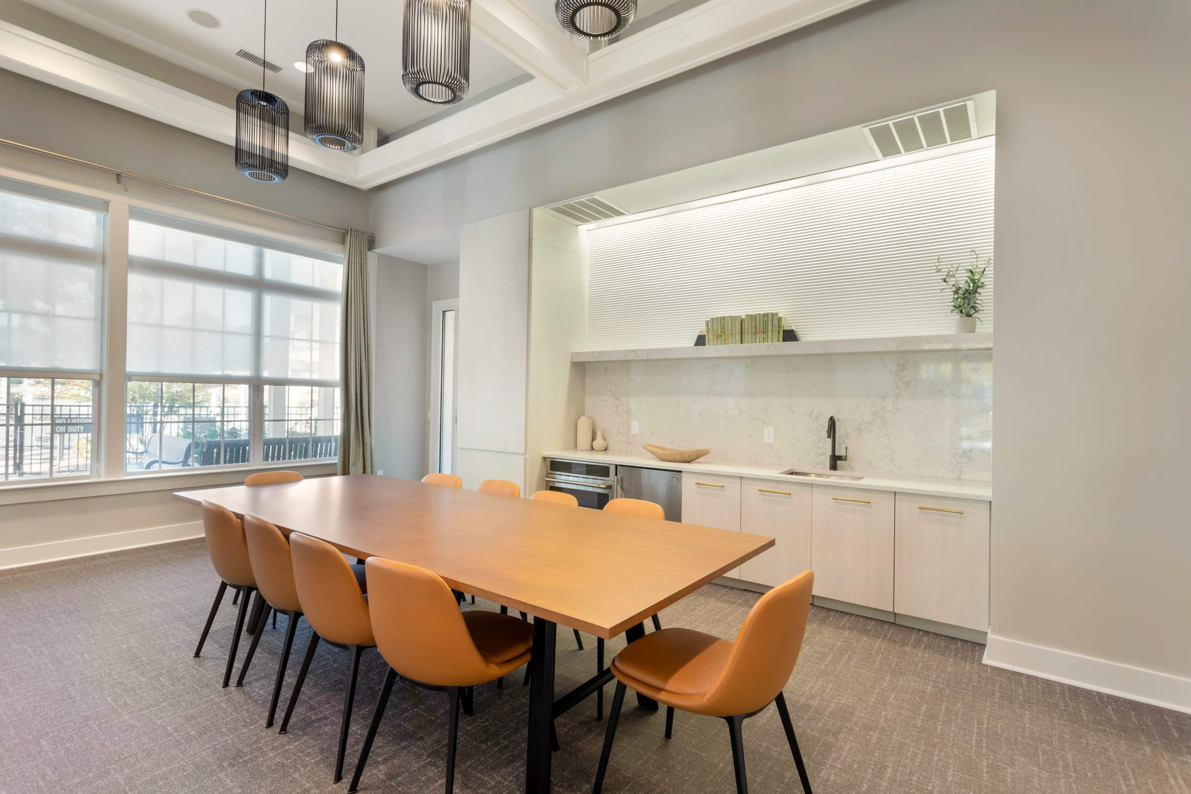 A modern conference room features a large wooden table surrounded by orange chairs, with a kitchenette area visible in the background.