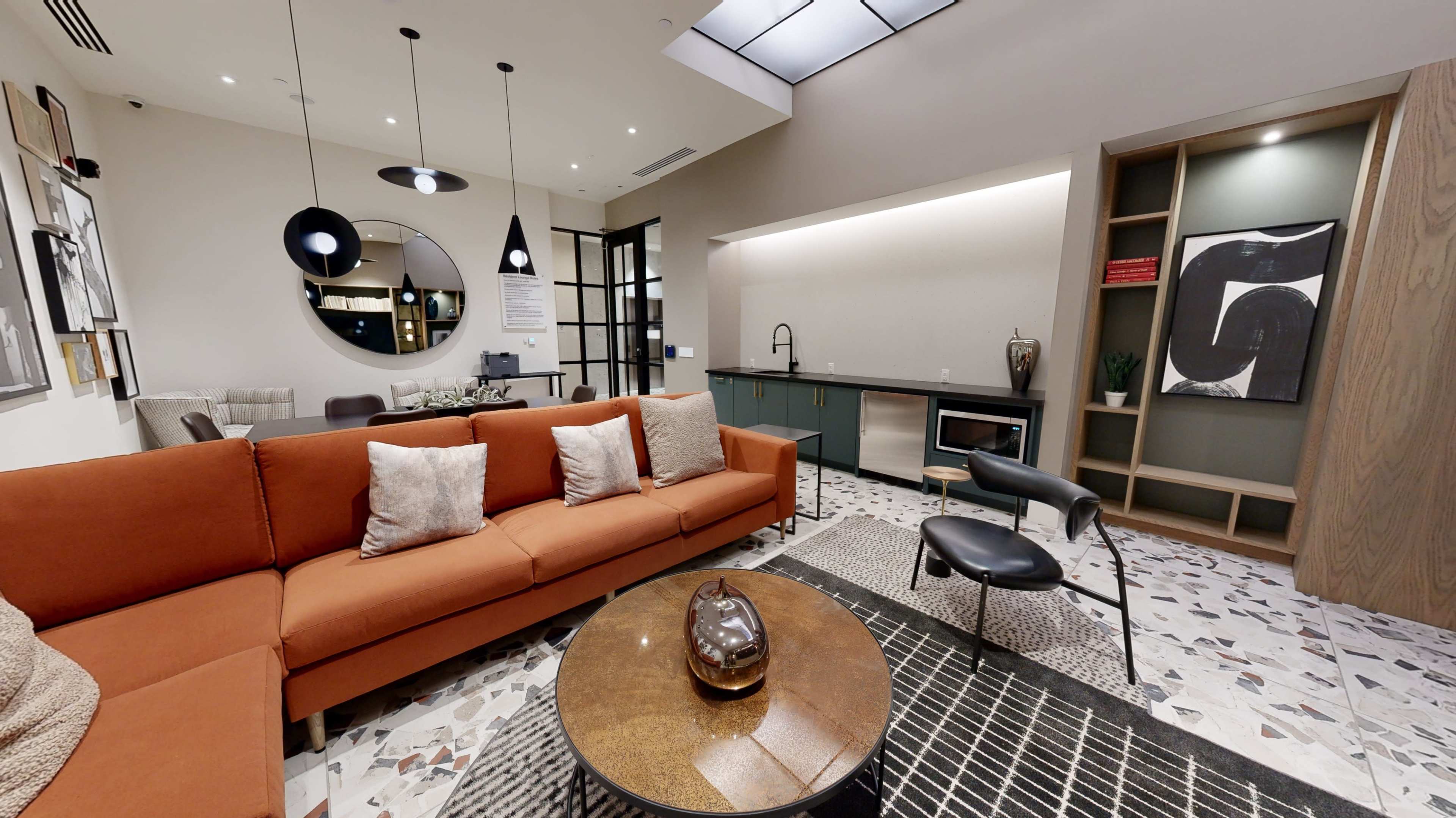 A modern living space featuring an orange sofa, a round mirror, and a coffee table on a patterned floor, with a kitchen area and shelving in the background.