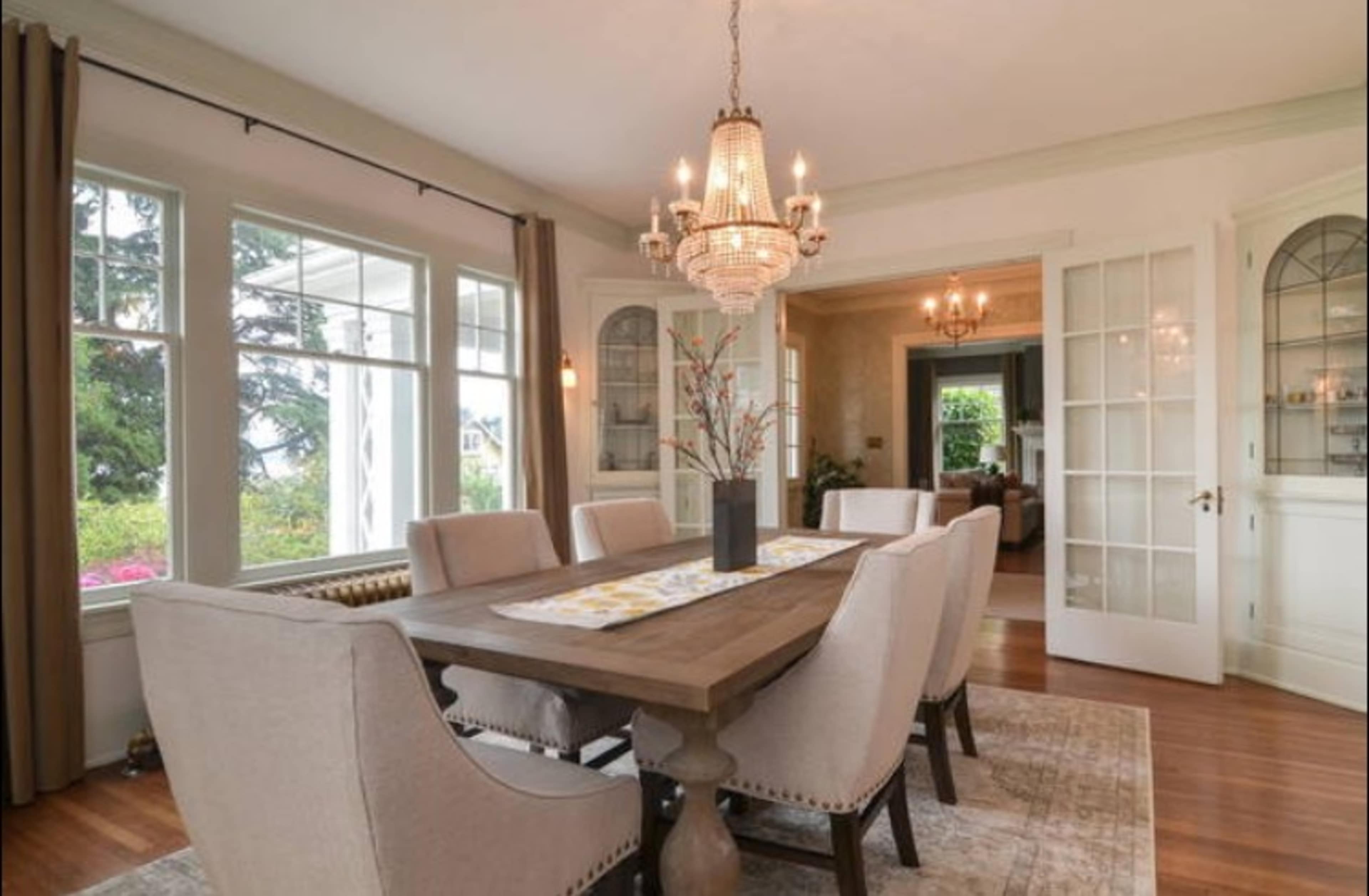 A dining room features a wooden table surrounded by upholstered chairs, with large windows allowing natural light and a chandelier above.