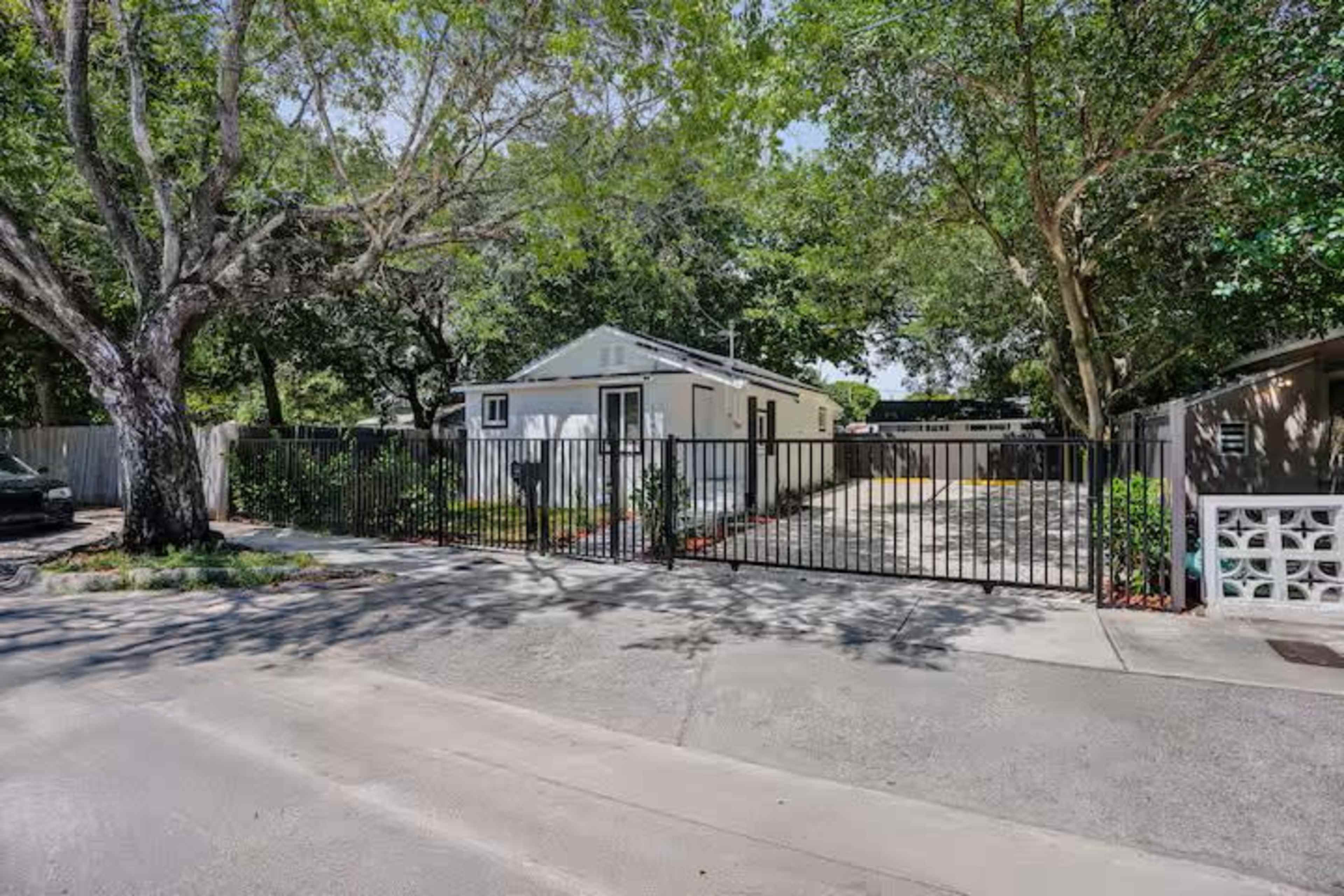The image shows a gated property with a single-story house framed by trees and a fenced area, situated along a street.