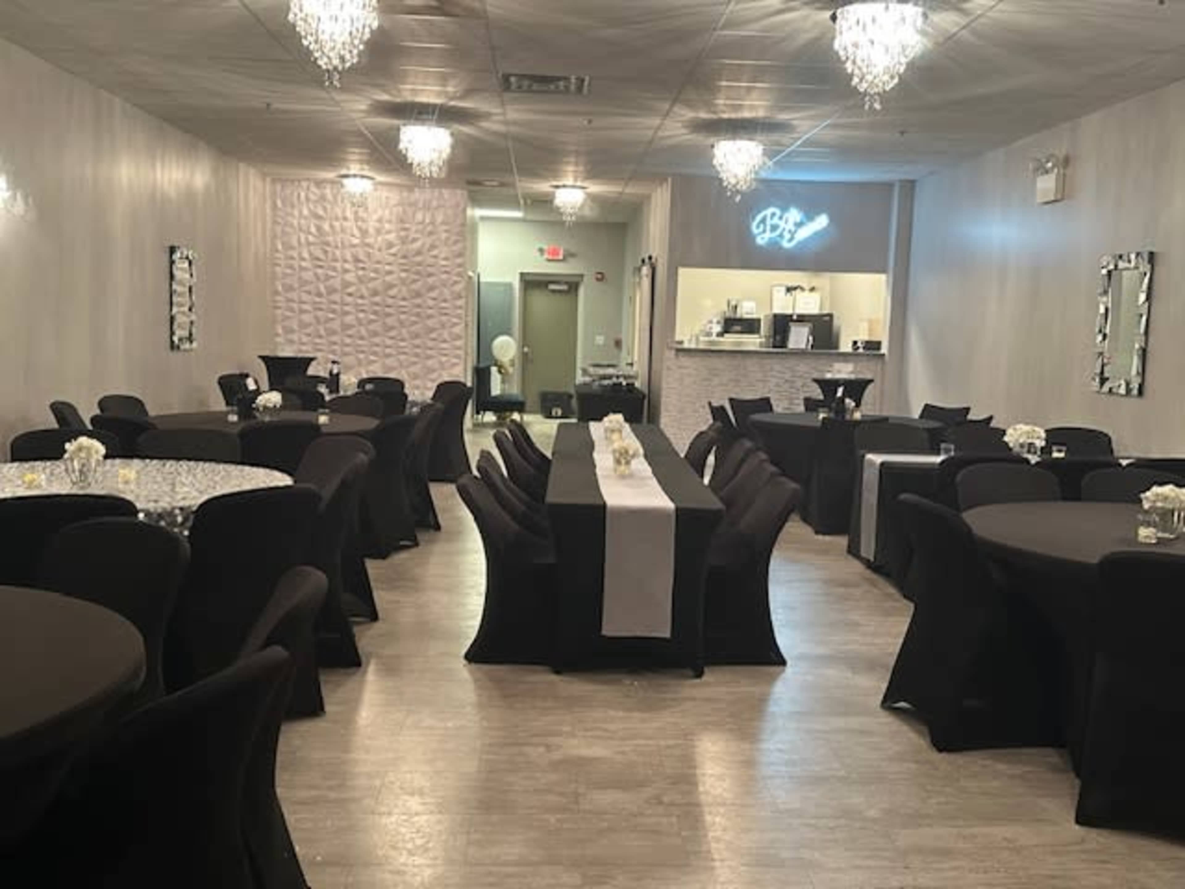 A spacious event venue with black clothed tables, white table runners, and elegant chandeliers overhead.