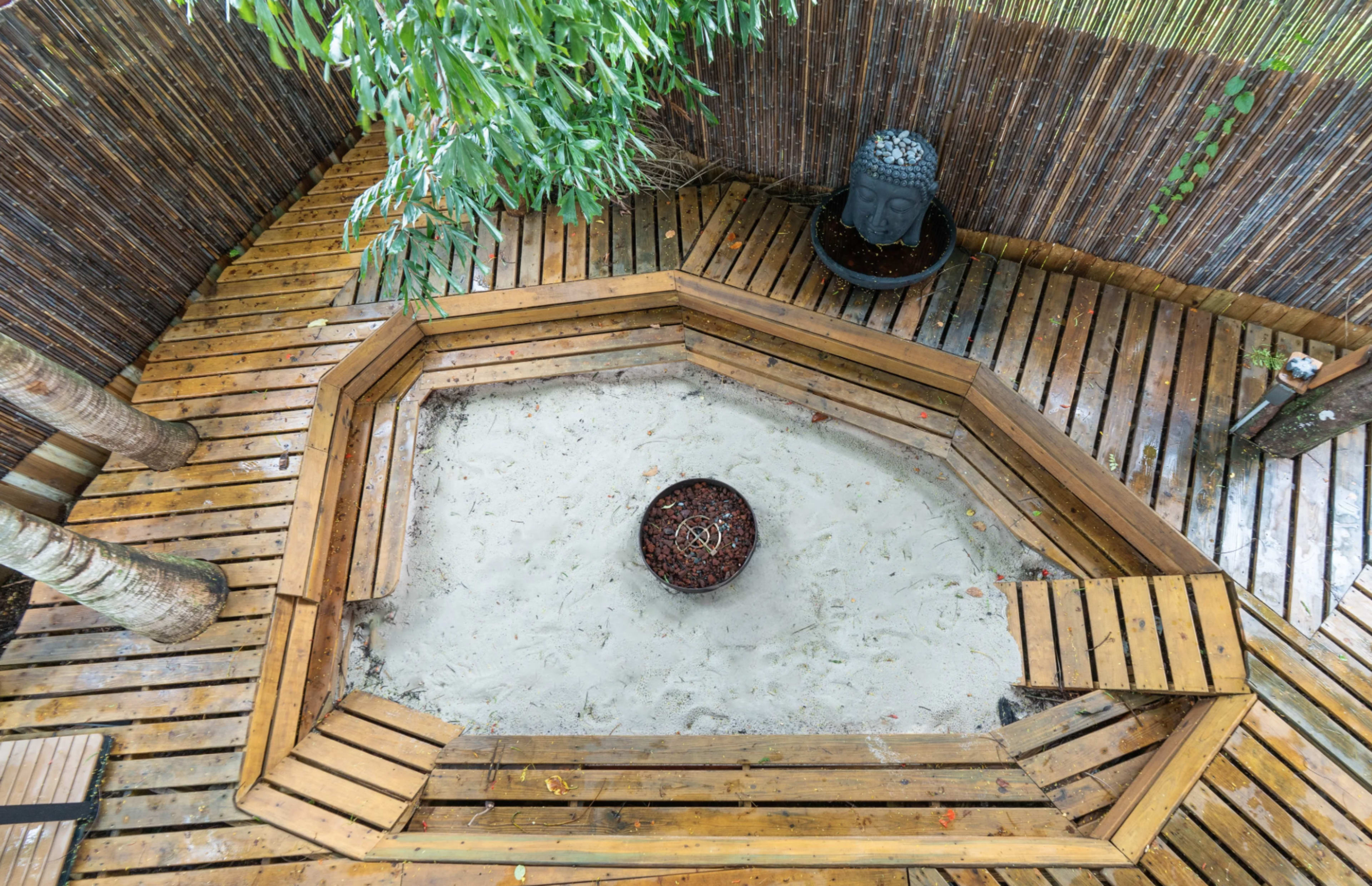The image shows a wooden octagonal seating area surrounding a sand pit, with a bamboo fence and greenery in the background.