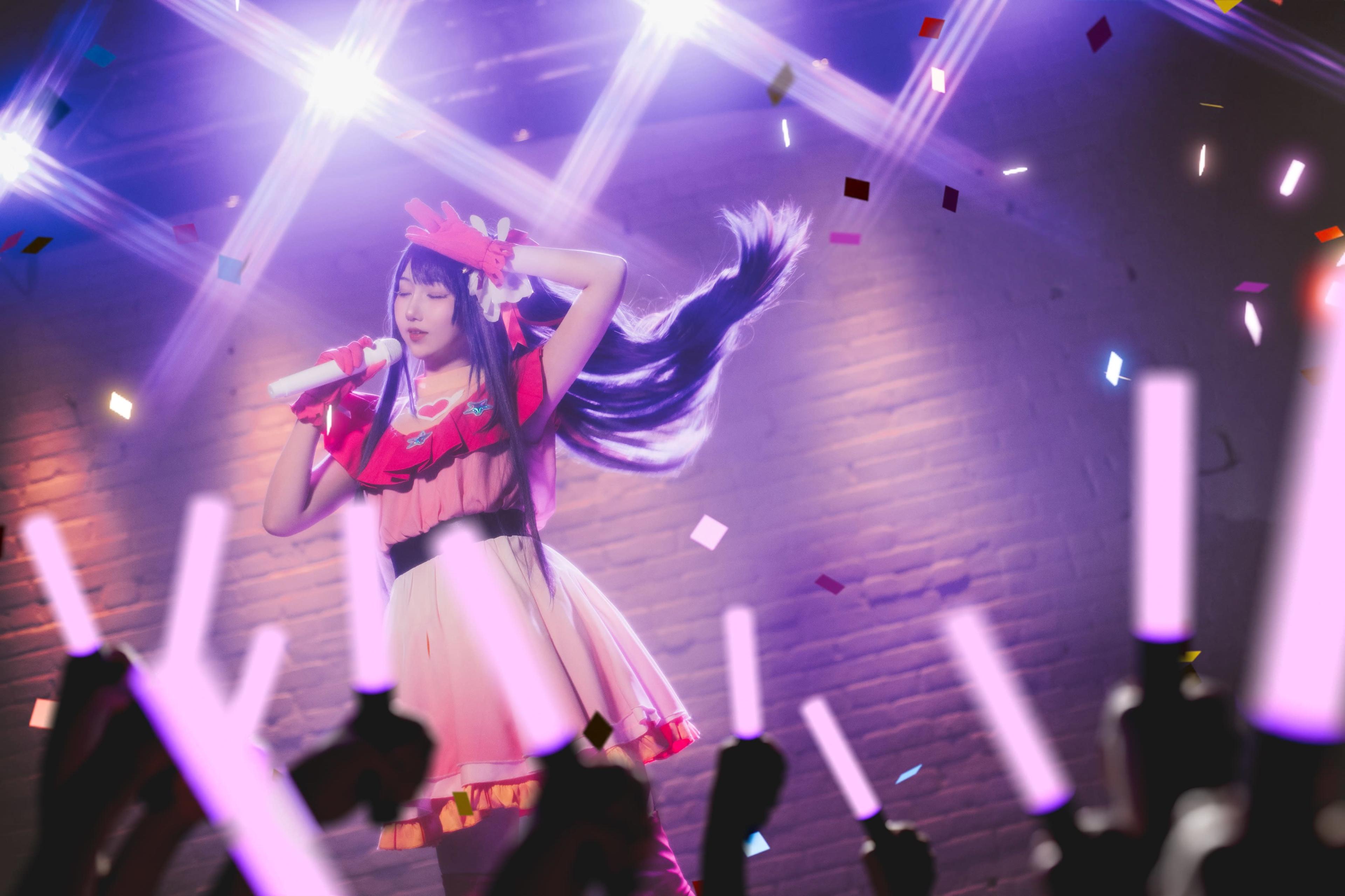 A performer dressed in a colorful costume sings on stage while fans wave light sticks in the air amidst a festive atmosphere.