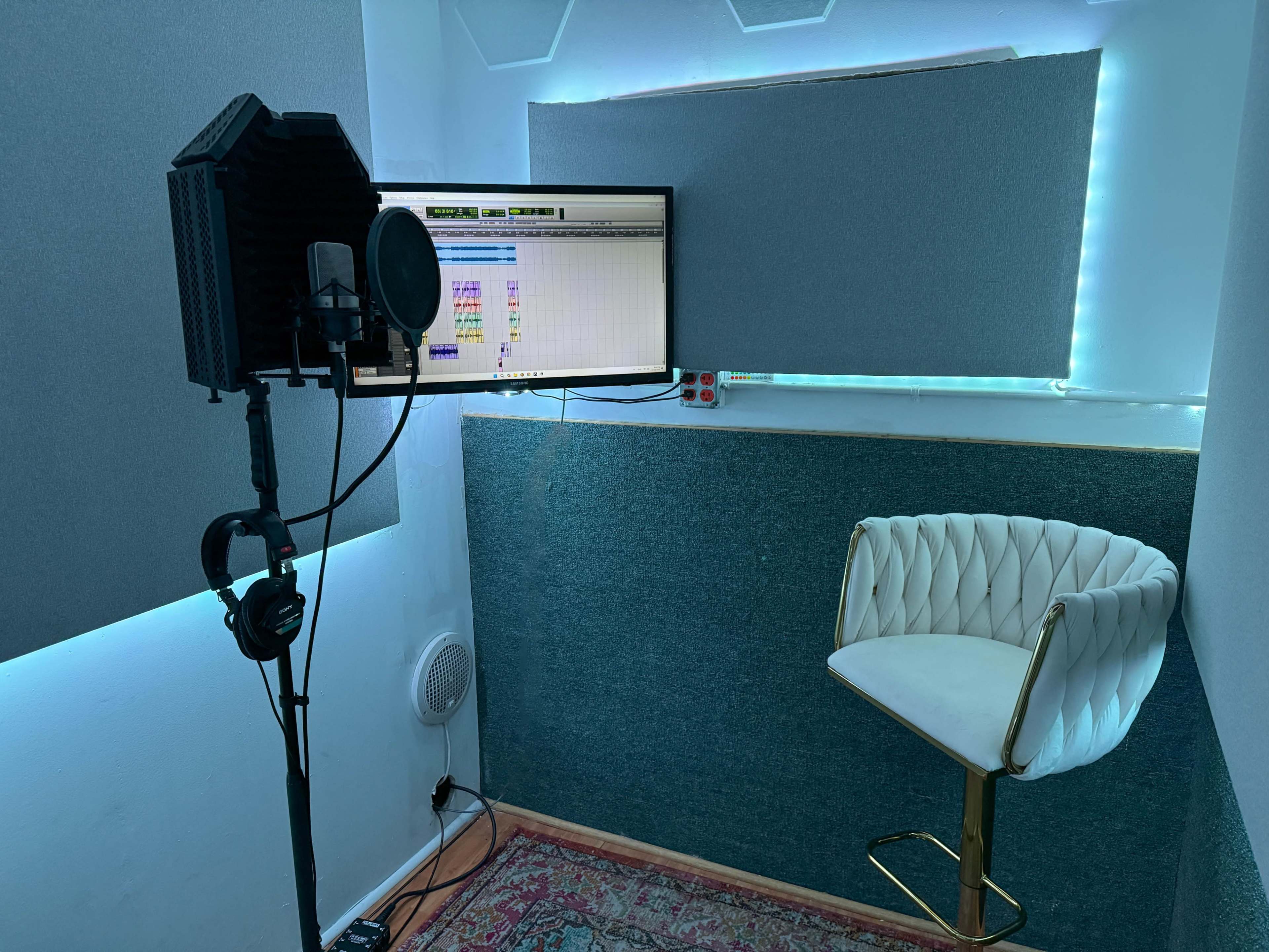 The image shows a sound recording studio with a microphone, a computer monitor displaying audio tracks, and a white bar stool in a small, padded room.