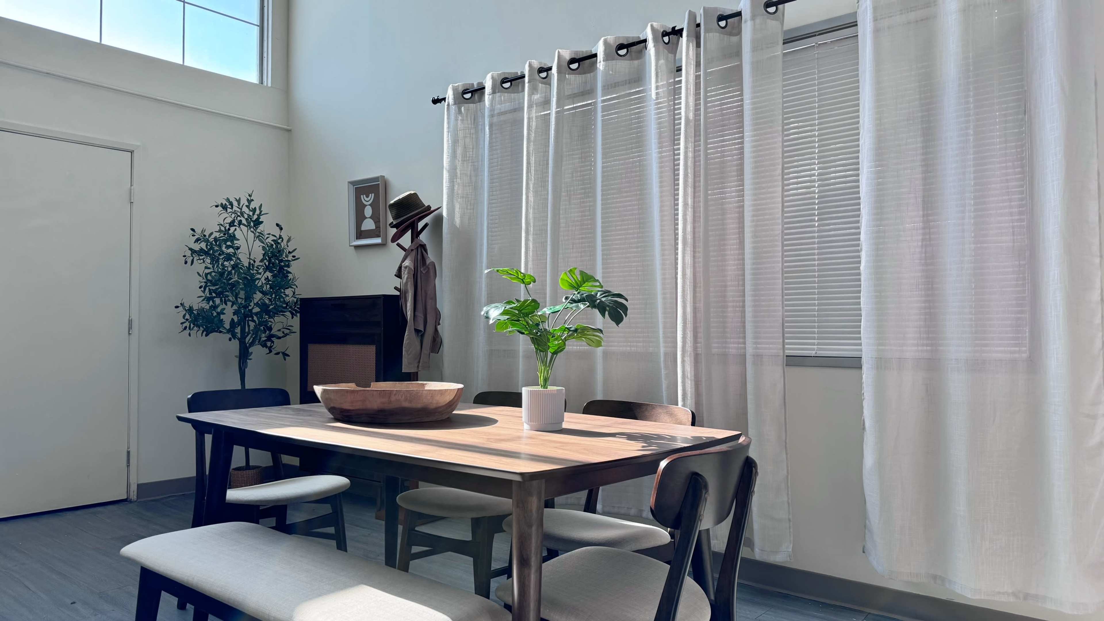 A wooden dining table with a bowl sits at the center of a well-lit room, surrounded by six chairs and featuring a potted plant by the window.