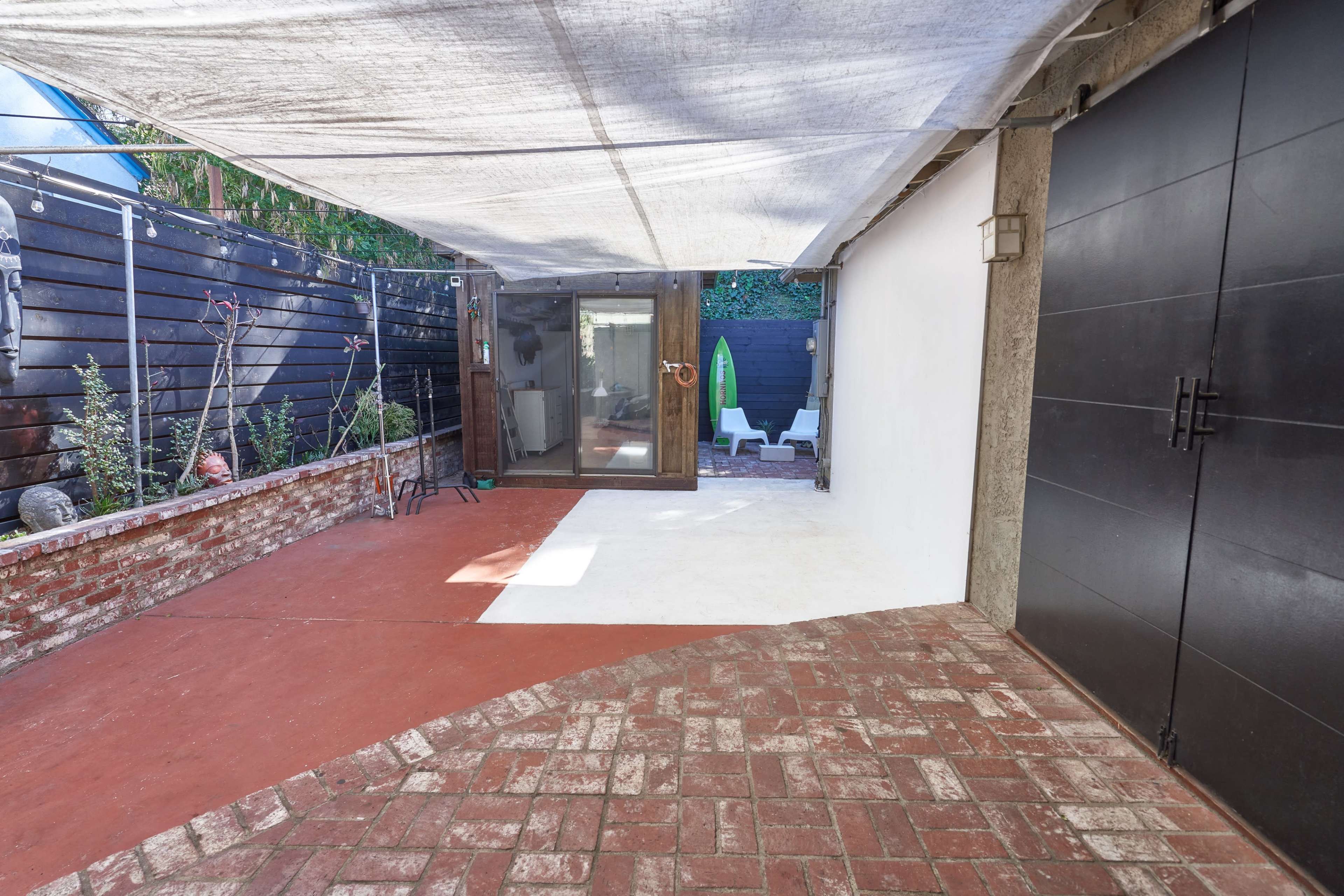 A covered outdoor space with a brick floor, featuring a glass door leading to a small enclosed area with seating and greenery.