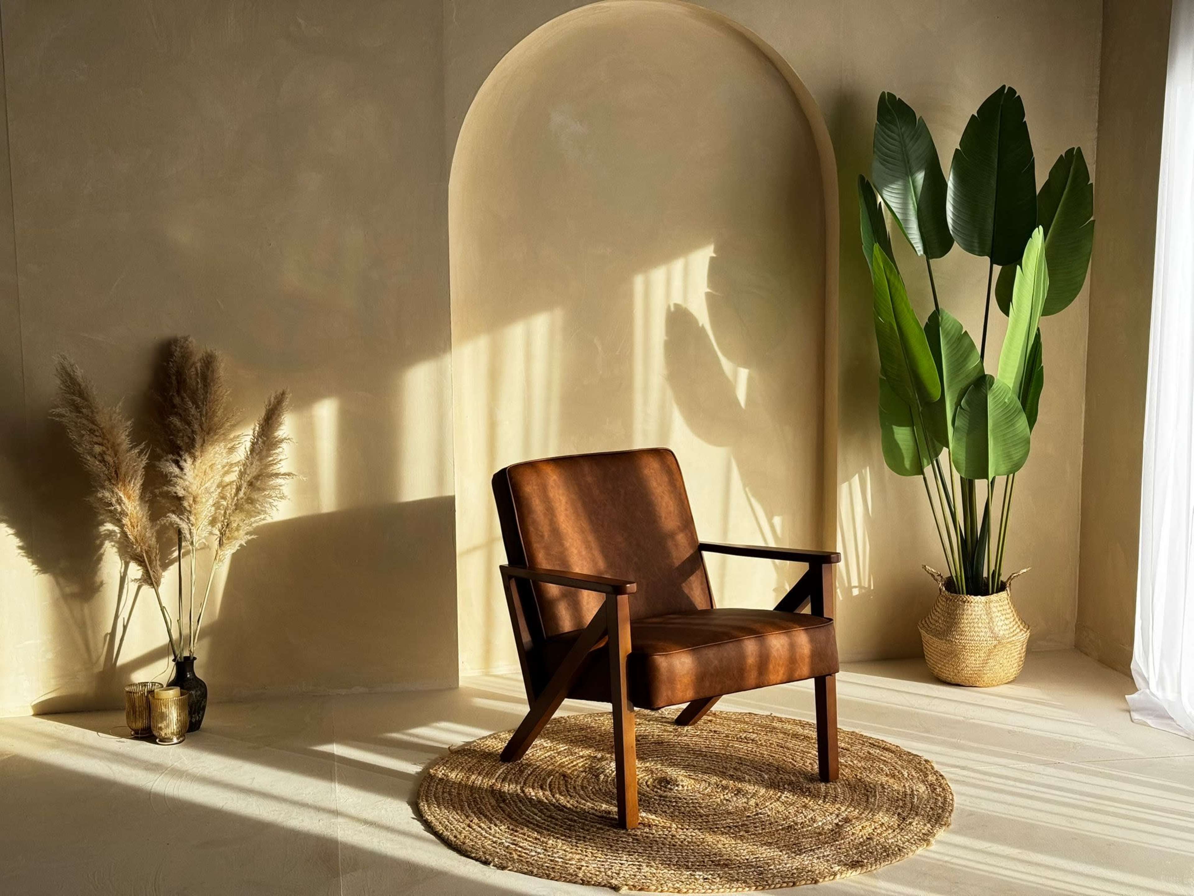 A brown armchair is positioned next to a large green plant and a decorative arrangement of dried grass in a softly lit, minimalist room.